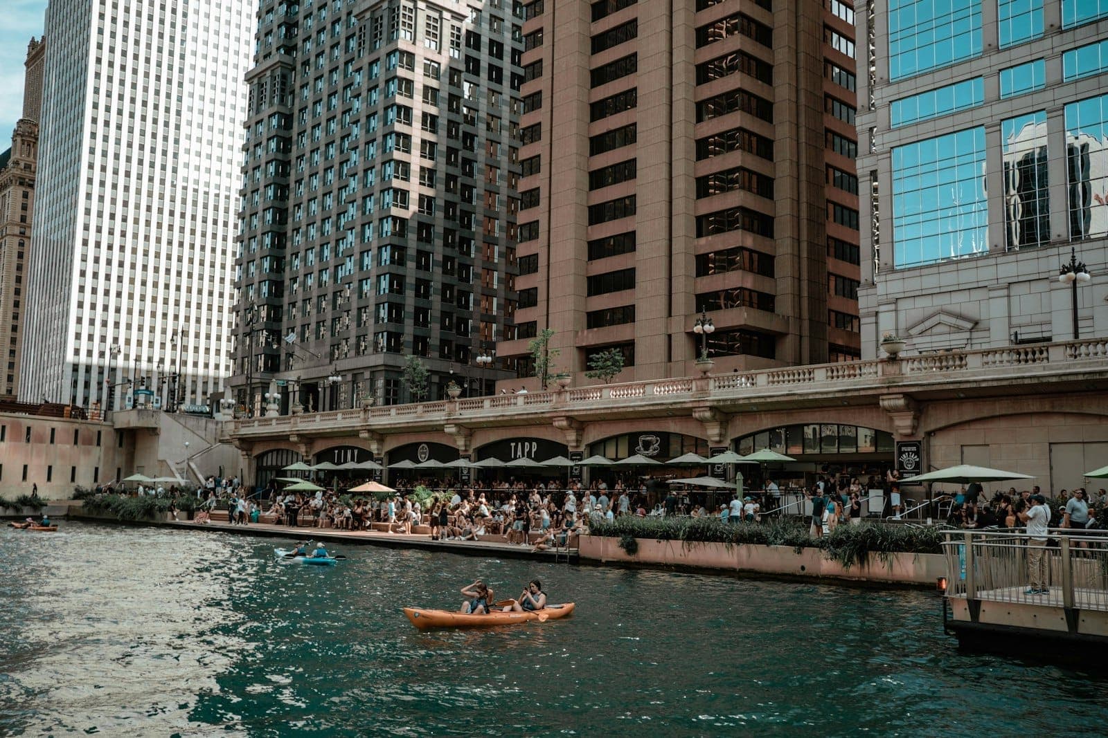 Chicago Riverwalk vào một ngày nắng đẹp với người dân dùng bữa tại các sân hiên ven sông và người chèo thuyền kayak trên sông, các tòa nhà cao tầng ở phía sau.