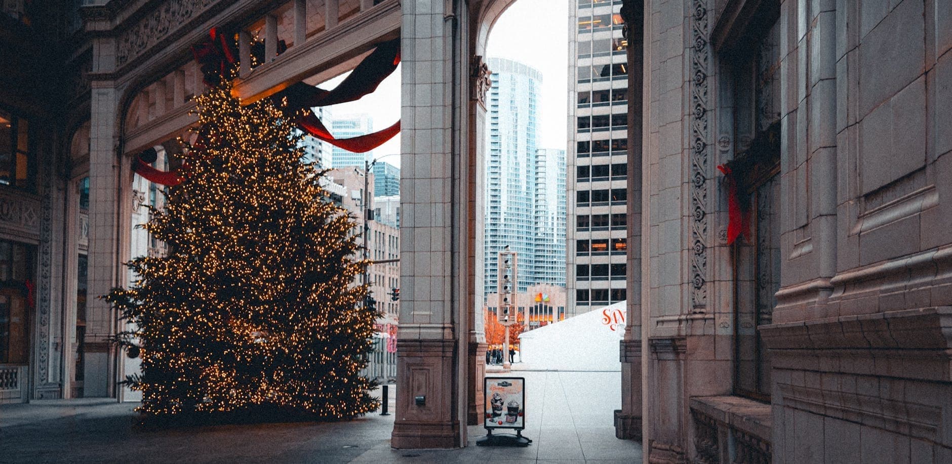 Large decorated Christmas tree with lights under an ornate archway in downtown Chicago surrounded by tall city buildings.