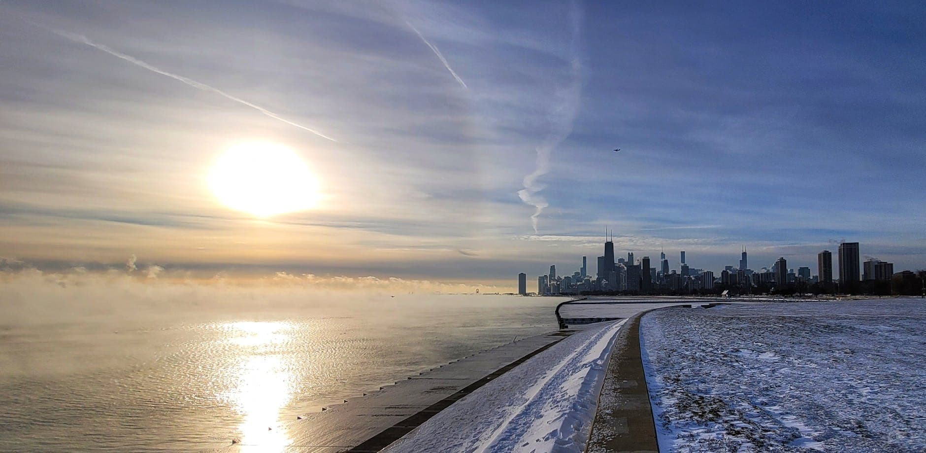 Snow-covered Chicago lakefront path with city skyline and mist rising off Lake Michigan in winter sunlight.