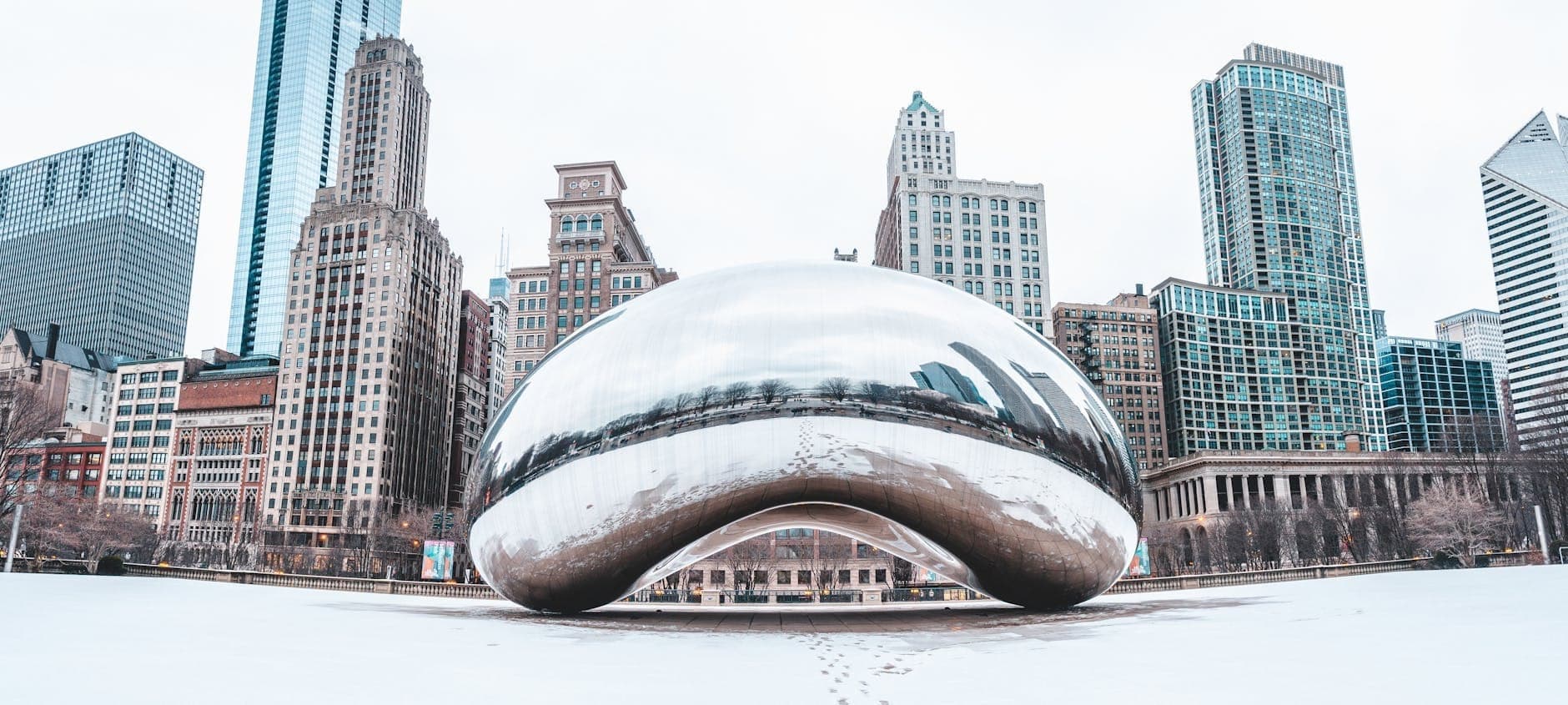 Wide view of Chicago’s Cloud Gate sculpture in Millennium Park, surrounded by snowy ground and city skyscrapers on a crisp winter day.