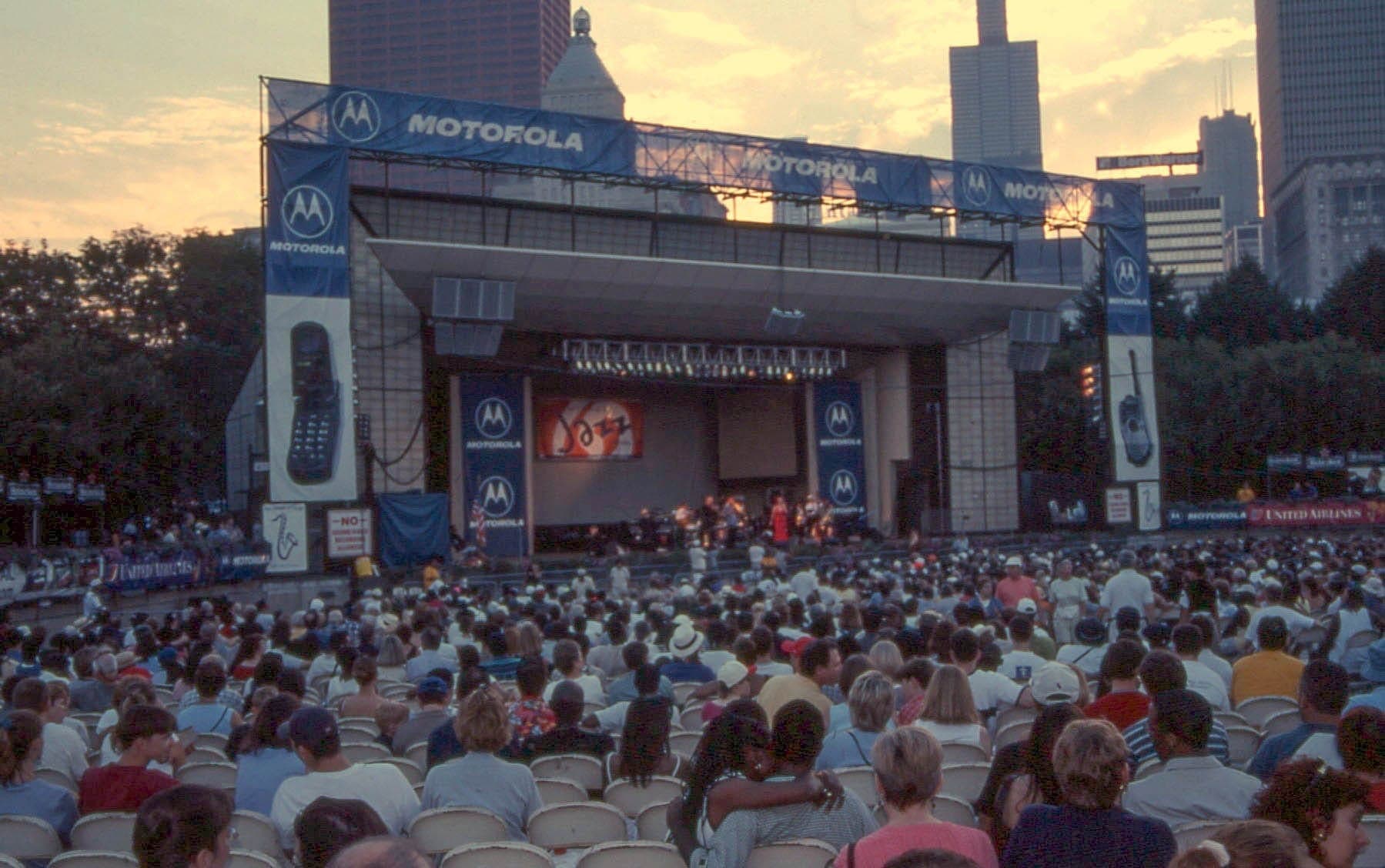 Große Menschenmenge genießt eine Live-Jazz-Darbietung auf der Freilichtbühne des Millennium Park in Chicago mit Wolkenkratzern bei Sonnenuntergang.