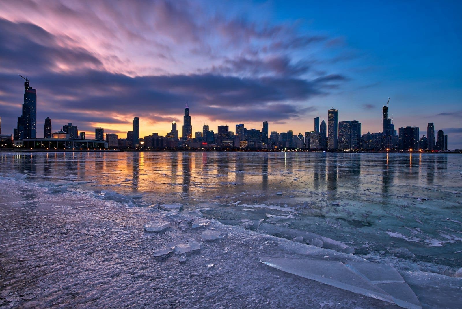 Icy Lake Michigan shoreline at sunset with Chicago skyline in the background, reflecting colorful clouds and city lights.
