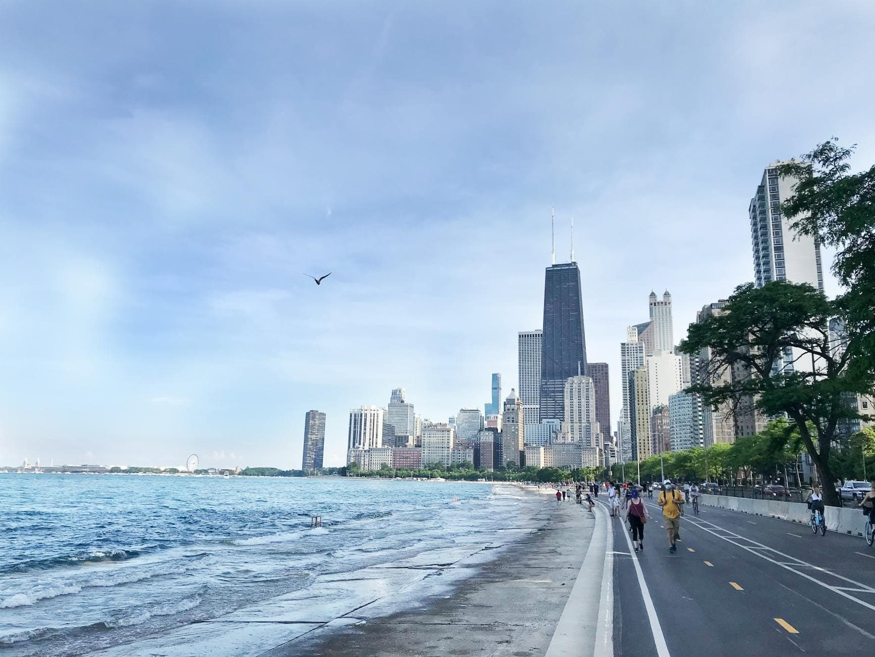 Paved trail along Lake Michigan with people walking and biking, Chicago skyline visible in background under blue sky.