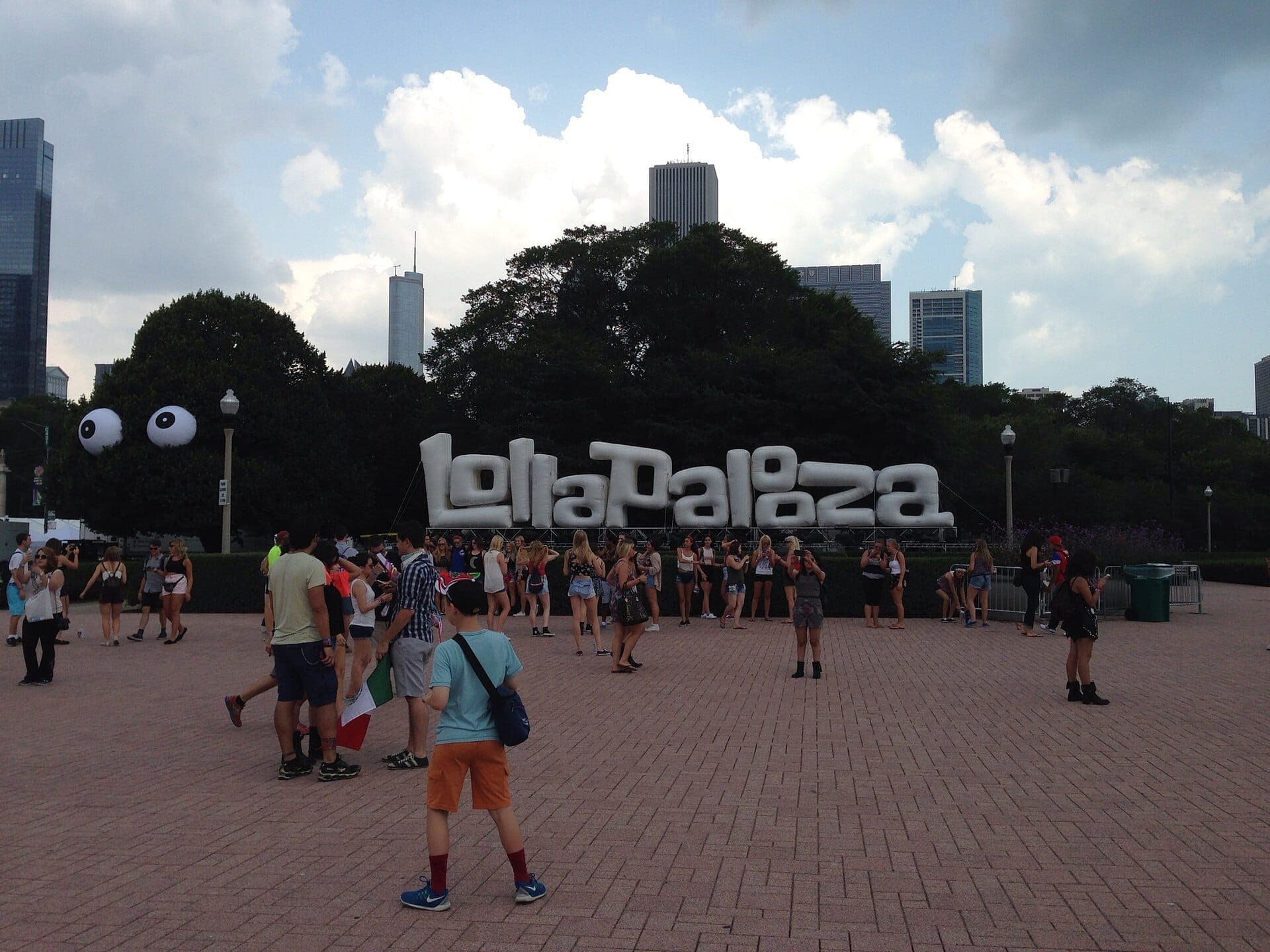 Du khách tập trung trước tấm biển Lollapalooza màu trắng lớn tại Grant Park, Chicago, với những tòa nhà chọc trời ở phía sau và những đám mây mùa hè phía trên.
