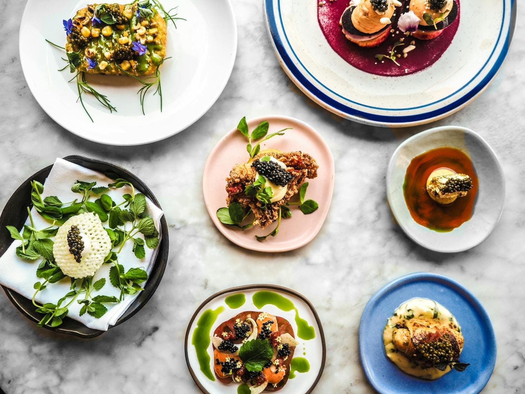 Assortment of beautifully plated gourmet dishes on colorful plates, seen from above on a marble table, evoking fine dining experience.