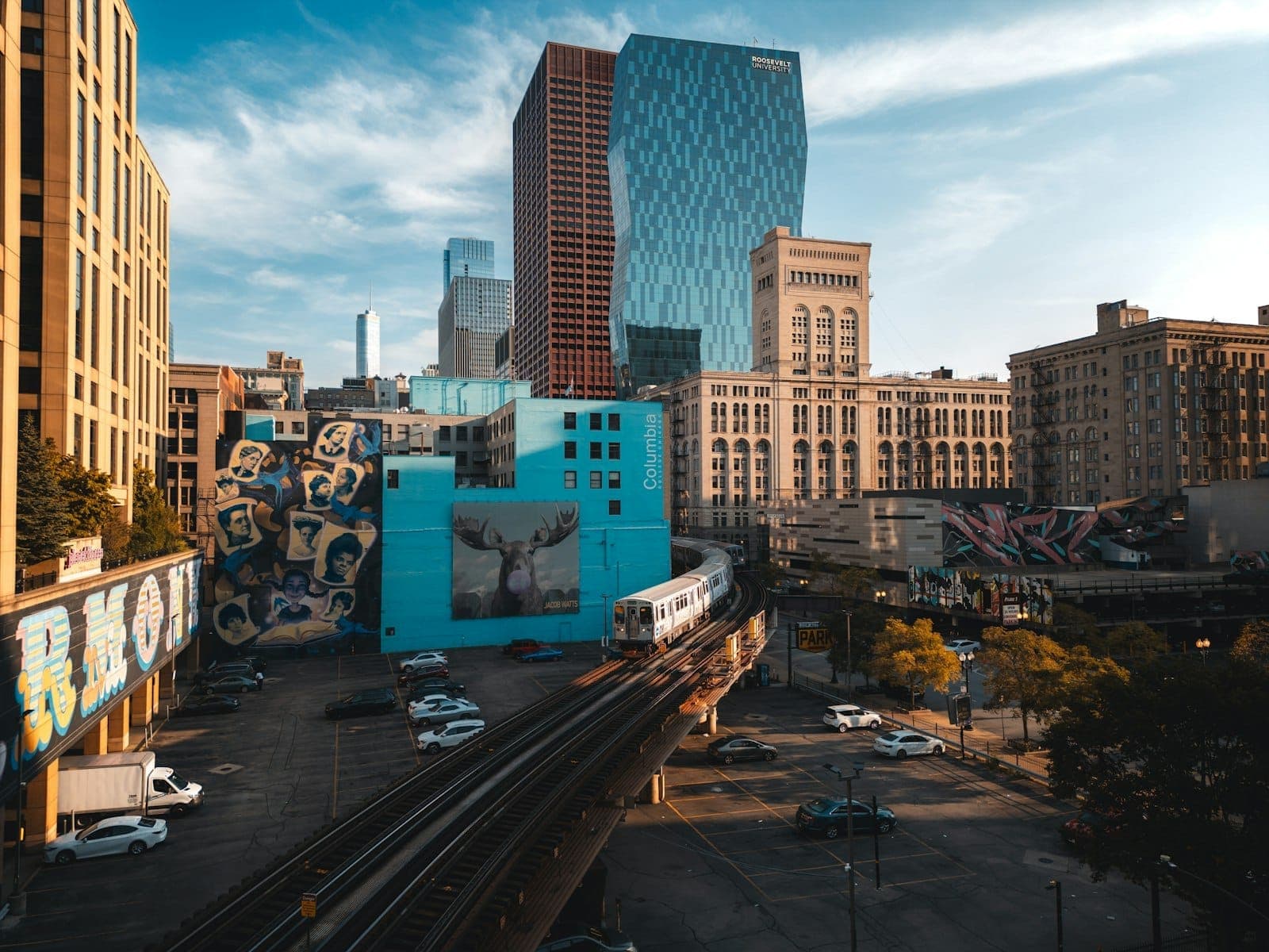 View of Chicago elevated train passing through South Side with tall modern buildings, urban murals, and city street in sunlight.