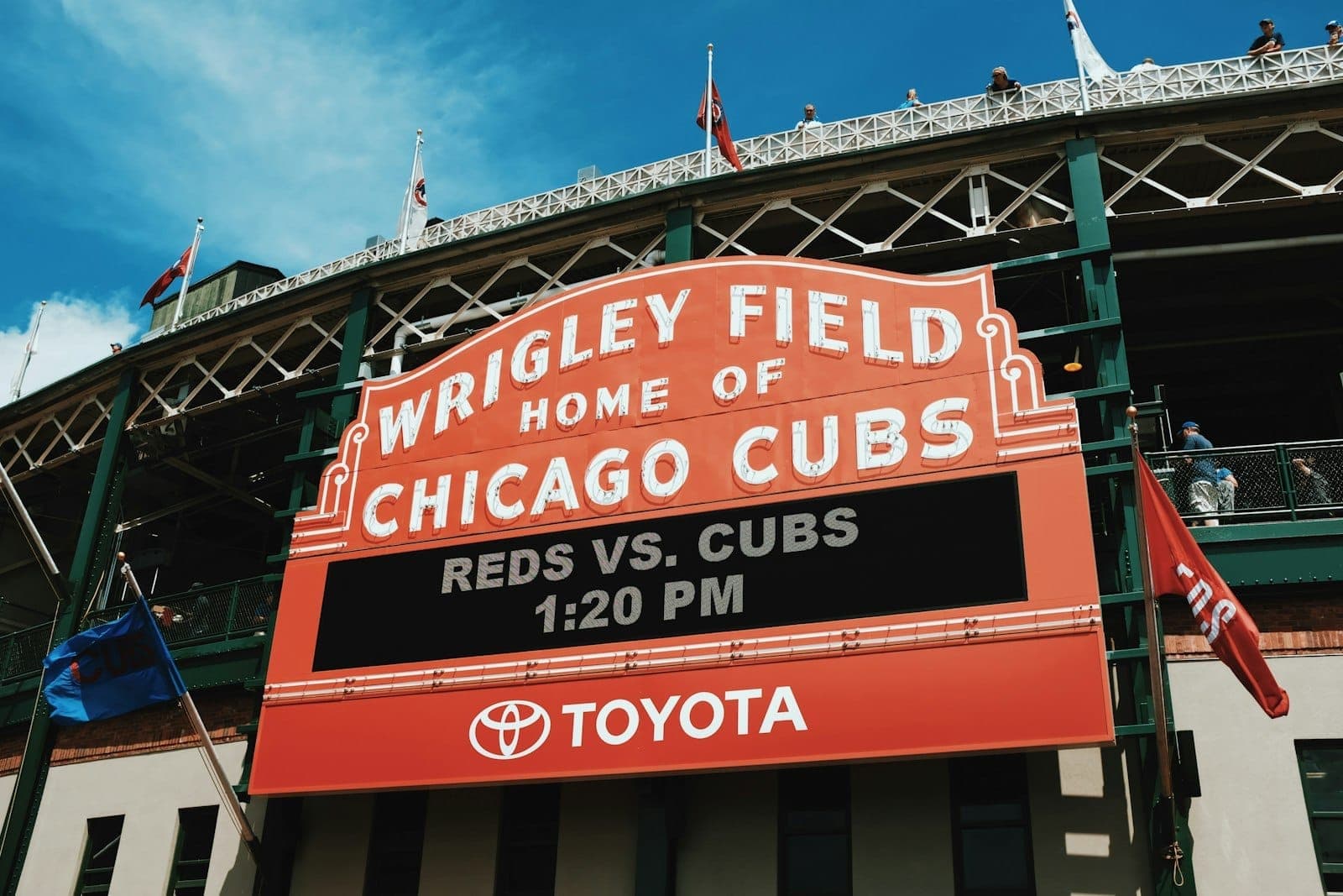 Wrigley Field’s iconic red marquee sign in Chicago, reading Wrigley Field Home of Chicago Cubs, with people visible on the roof above.