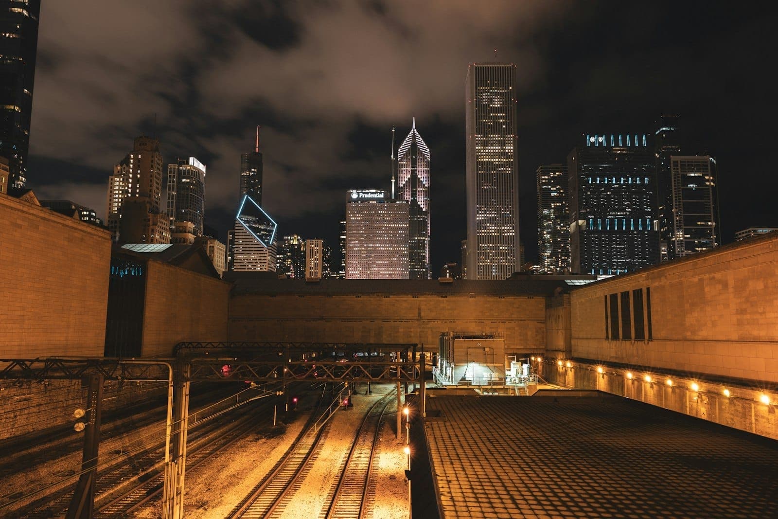 Night view of Chicago skyscrapers from a rooftop deck, with glowing city lights and dramatic clouds above.