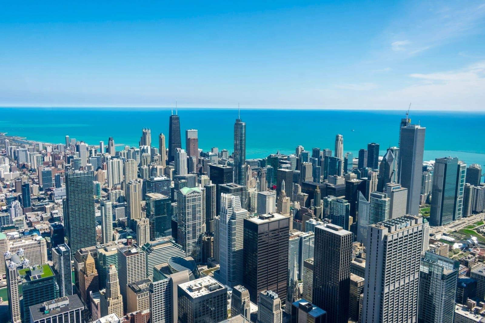 Bright, clear daytime aerial view of downtown Chicago skyscrapers with Lake Michigan in the background, seen from a high observation deck perspective.