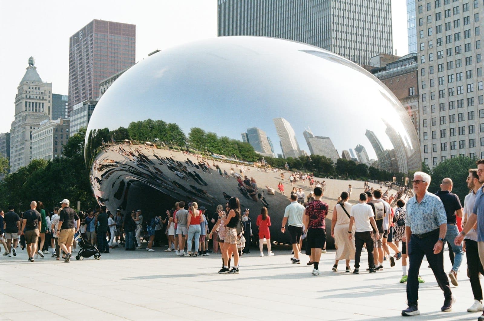 Crowds of people gather at Cloud Gate (The Bean) in Millennium Park with Chicago skyscrapers in the background on a sunny day.