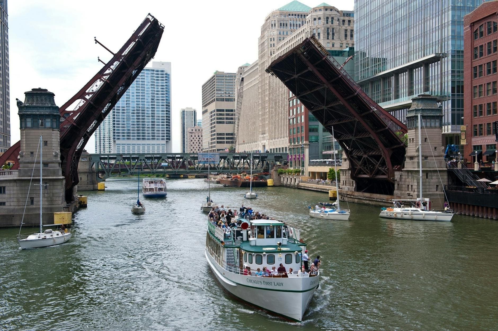 A riverboat full of people passes under a raised bascule bridge on the Chicago River, surrounded by skyscrapers and city activity.