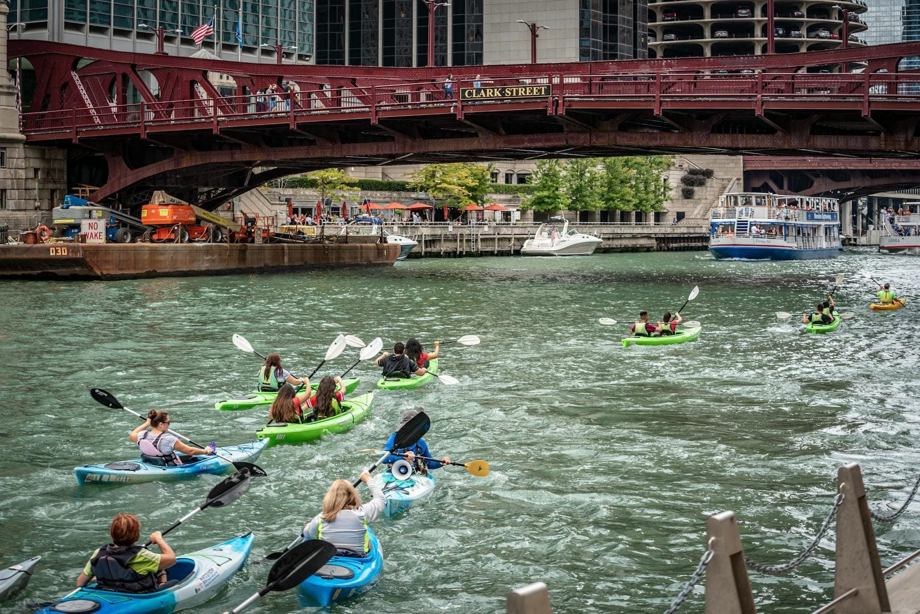 Group of kayakers paddling on the Chicago River under a red bridge with skyscrapers and a sightseeing boat in the background.