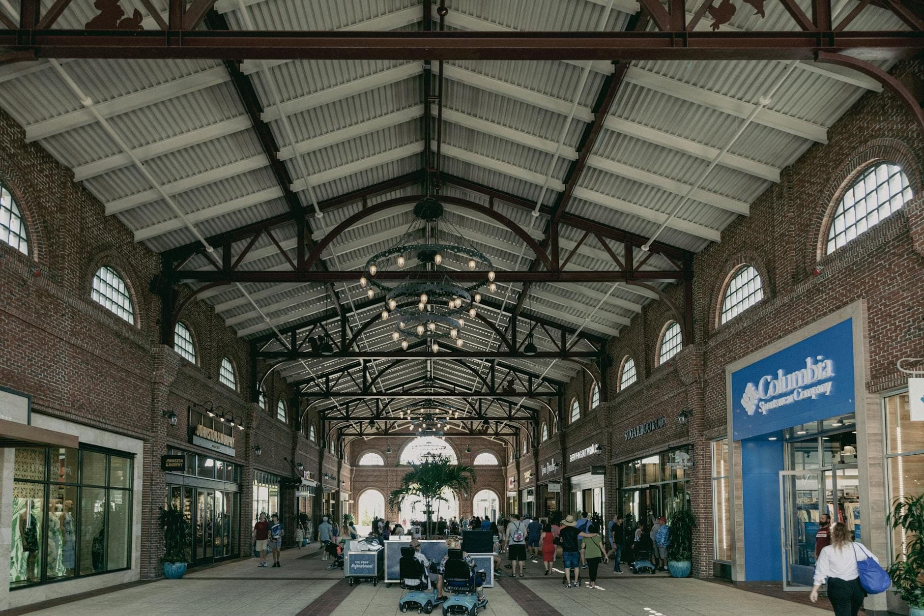 Interior view of an outlet-style shopping center with brick walls, glass storefronts, and shoppers walking beneath a high, industrial-style ceiling.