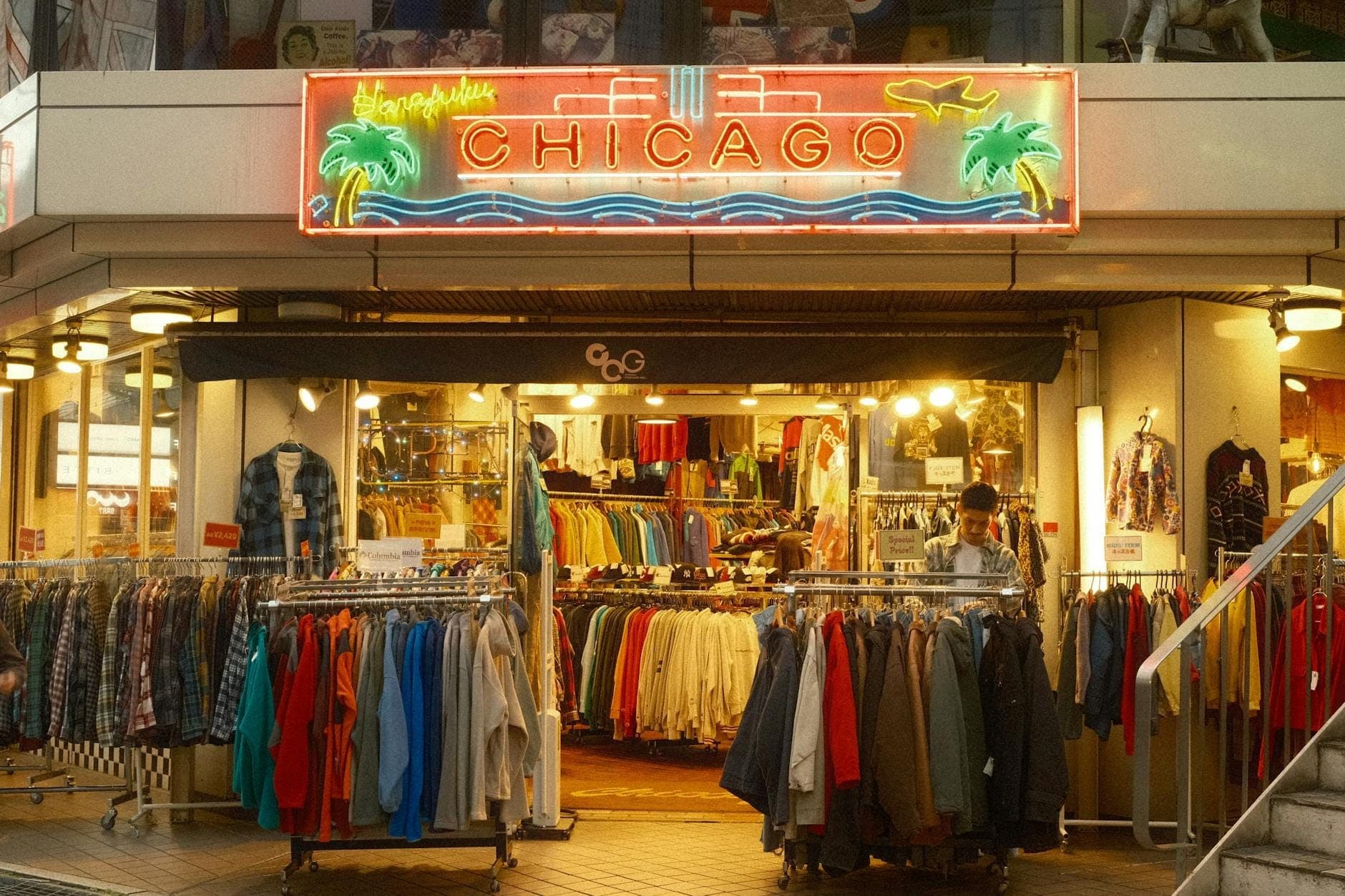 Storefront with racks of clothes outside, neon sign reading 'Chicago' above entrance, and a person browsing inside.