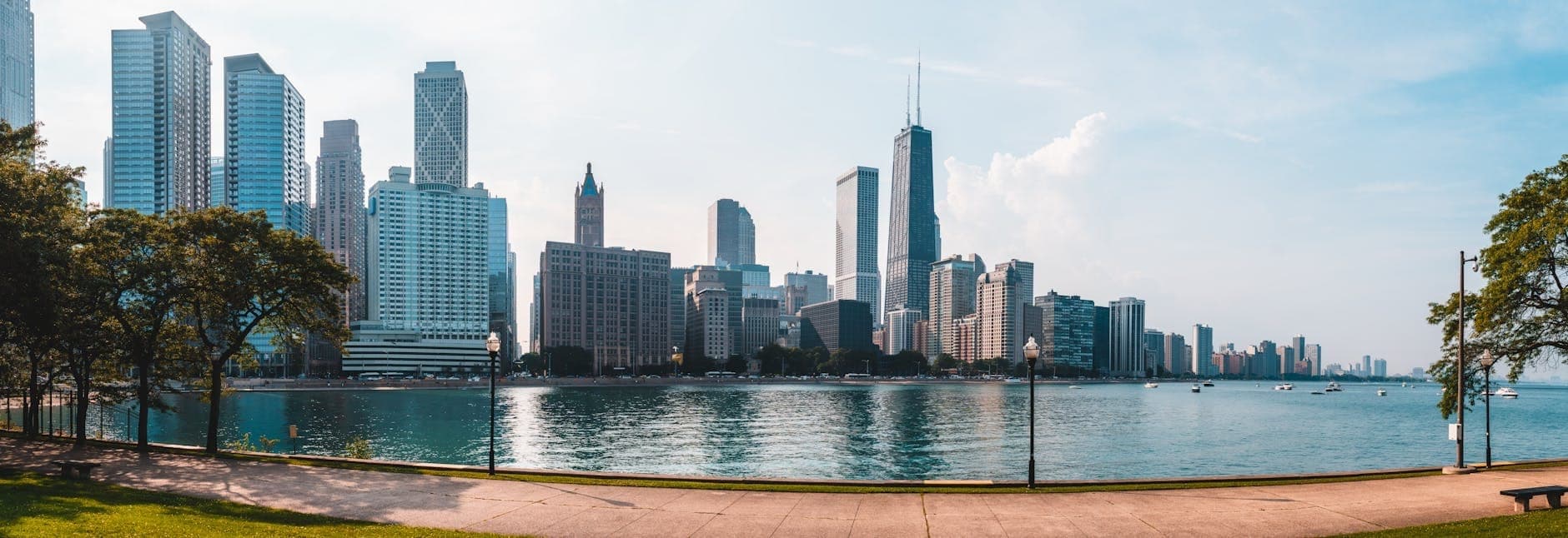 Panoramic view of the Chicago skyline from the waterfront park on a sunny day, featuring tall skyscrapers, clear blue sky, and lakeside walking paths.