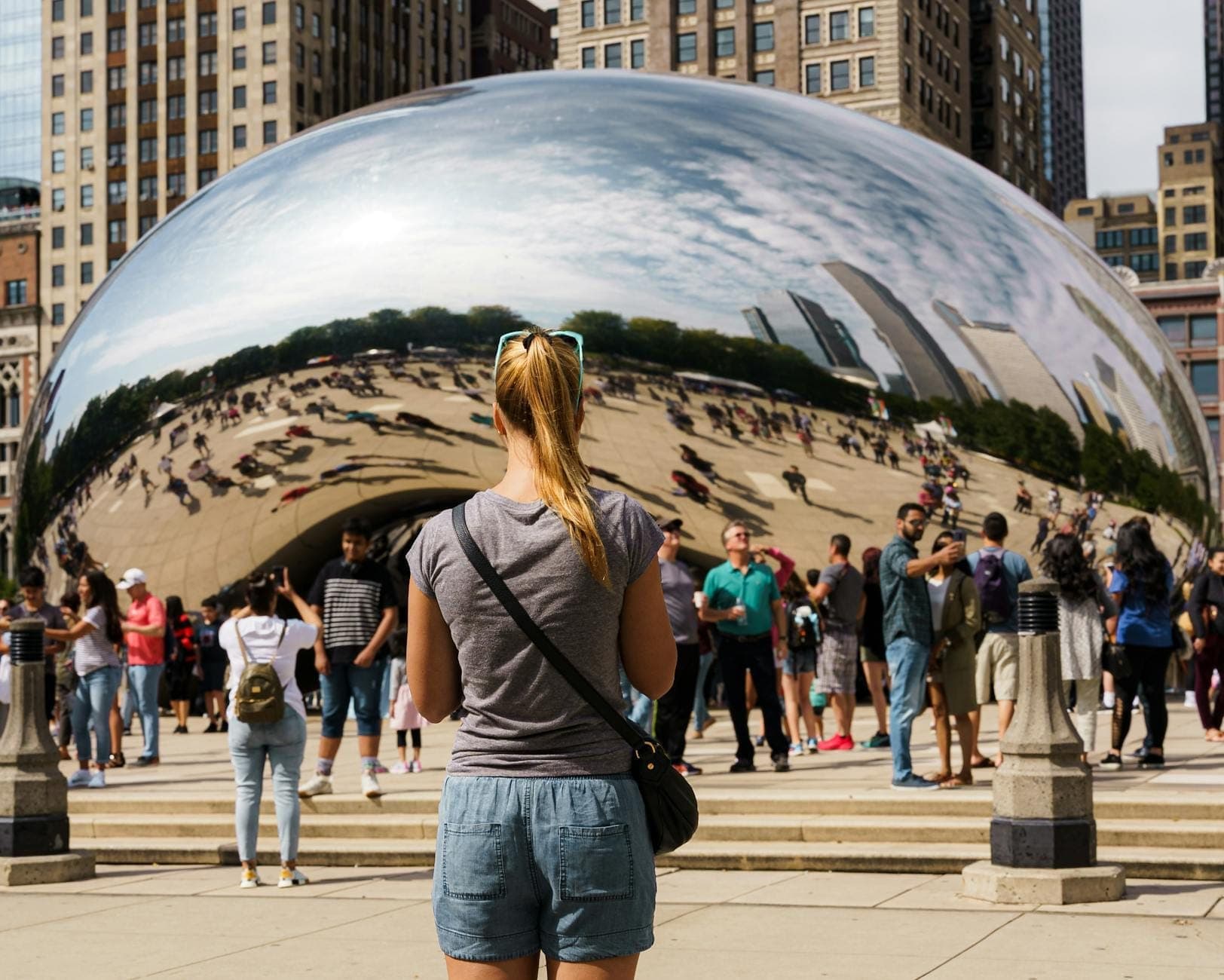 A solo traveler faces Chicago’s Cloud Gate (The Bean) amid a lively crowd, surrounded by downtown skyscrapers on a sunny day.
