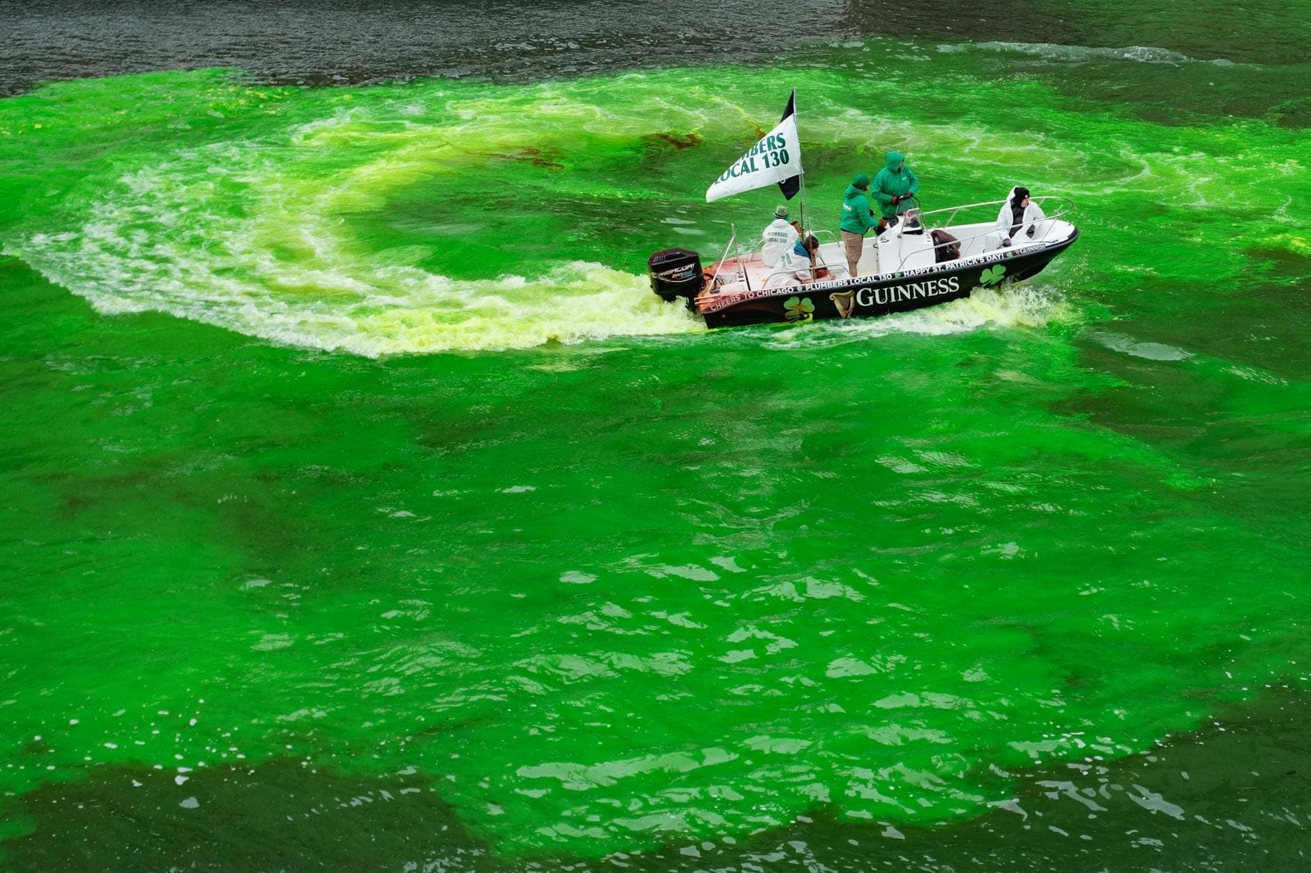 Small boat with people in costume and flags sailing through bright green dyed Chicago River during St. Patrick’s Day celebration.