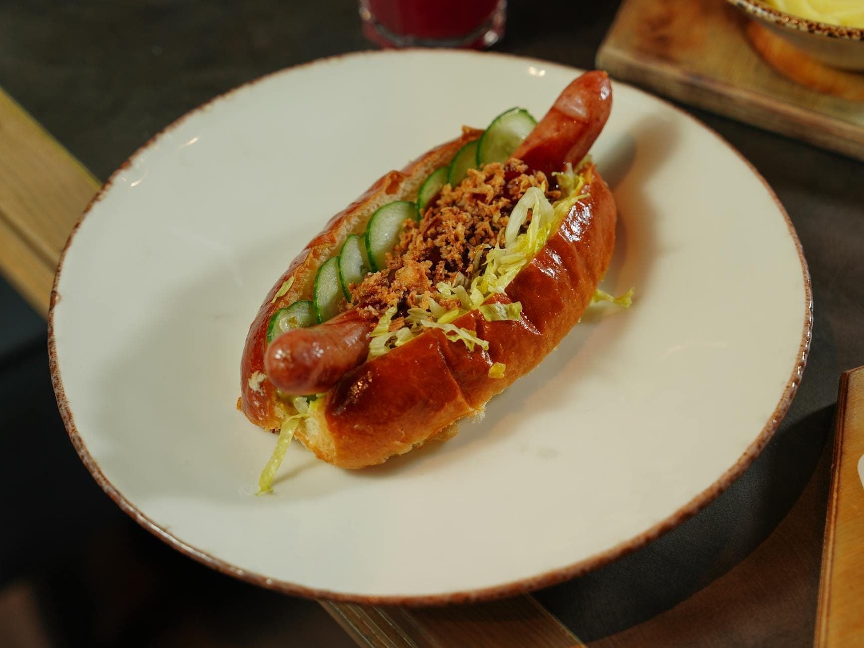 A Chicago style hot dog topped with sliced cucumber, shredded lettuce, and crispy bits on a white plate in soft indoor lighting.