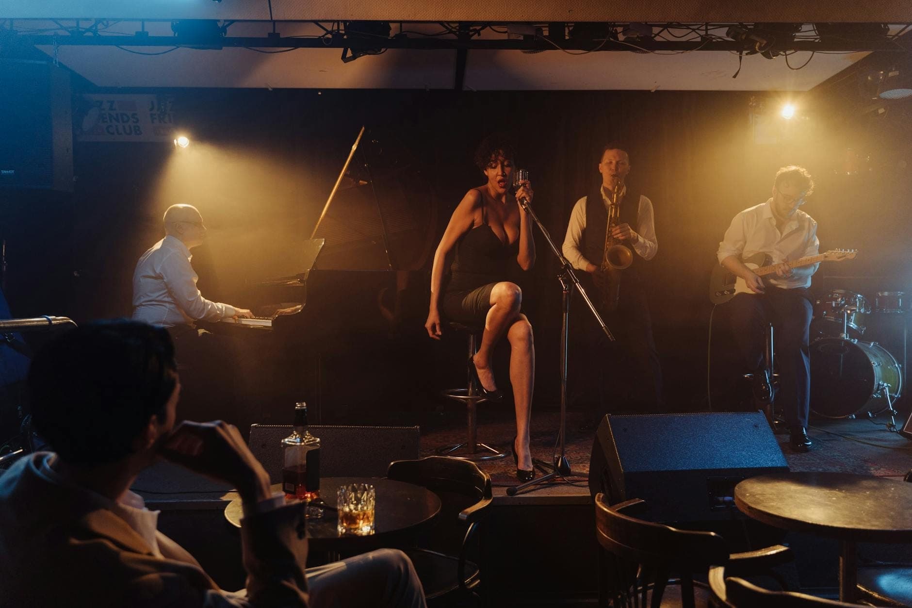 Jazz-Quartett auf der Bühne eines intimen, schwach beleuchteten Clubs, aus der Perspektive des Publikums mit Getränken auf kleinen Tischen.