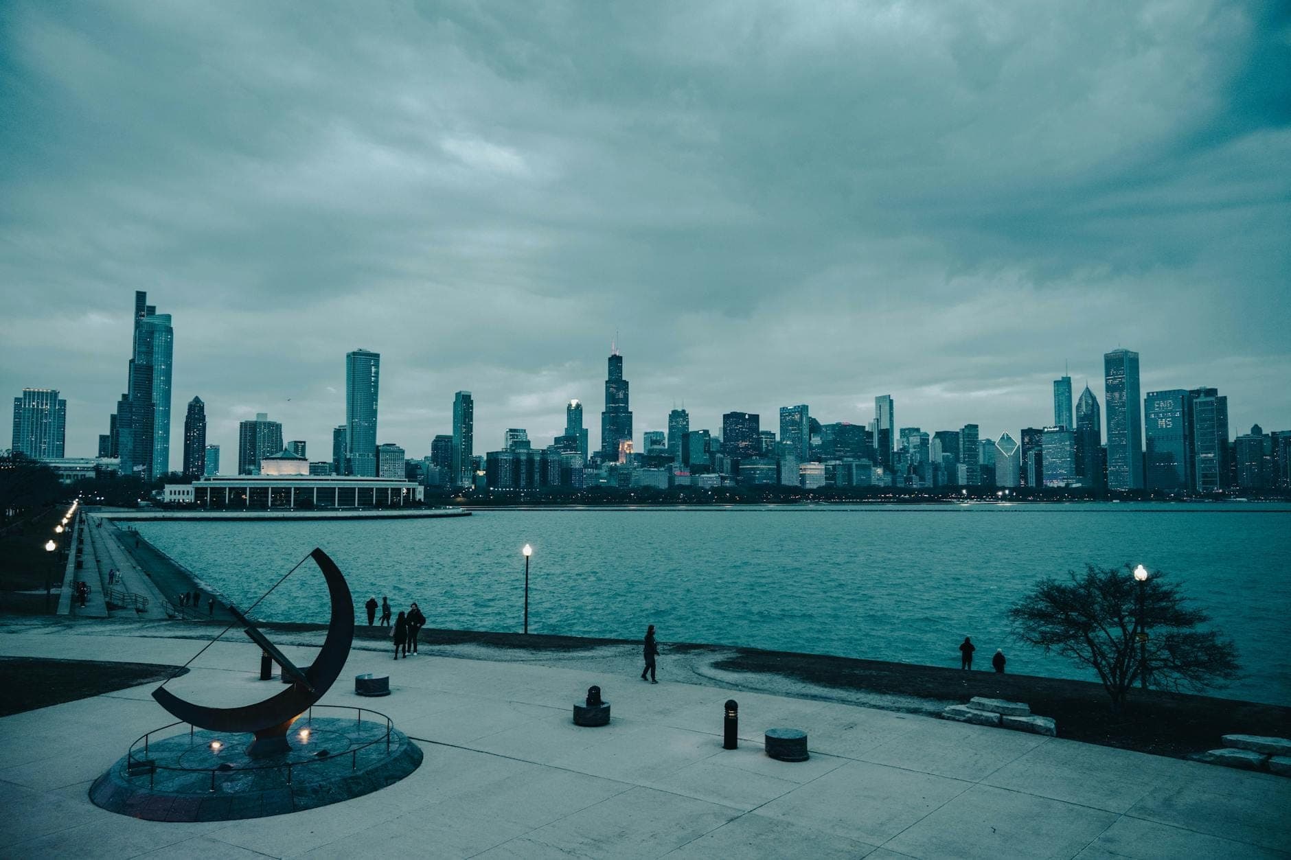 View of the Chicago skyline from a lakeside pathway with sculptures and people, overlooking Lake Michigan on a cloudy day.