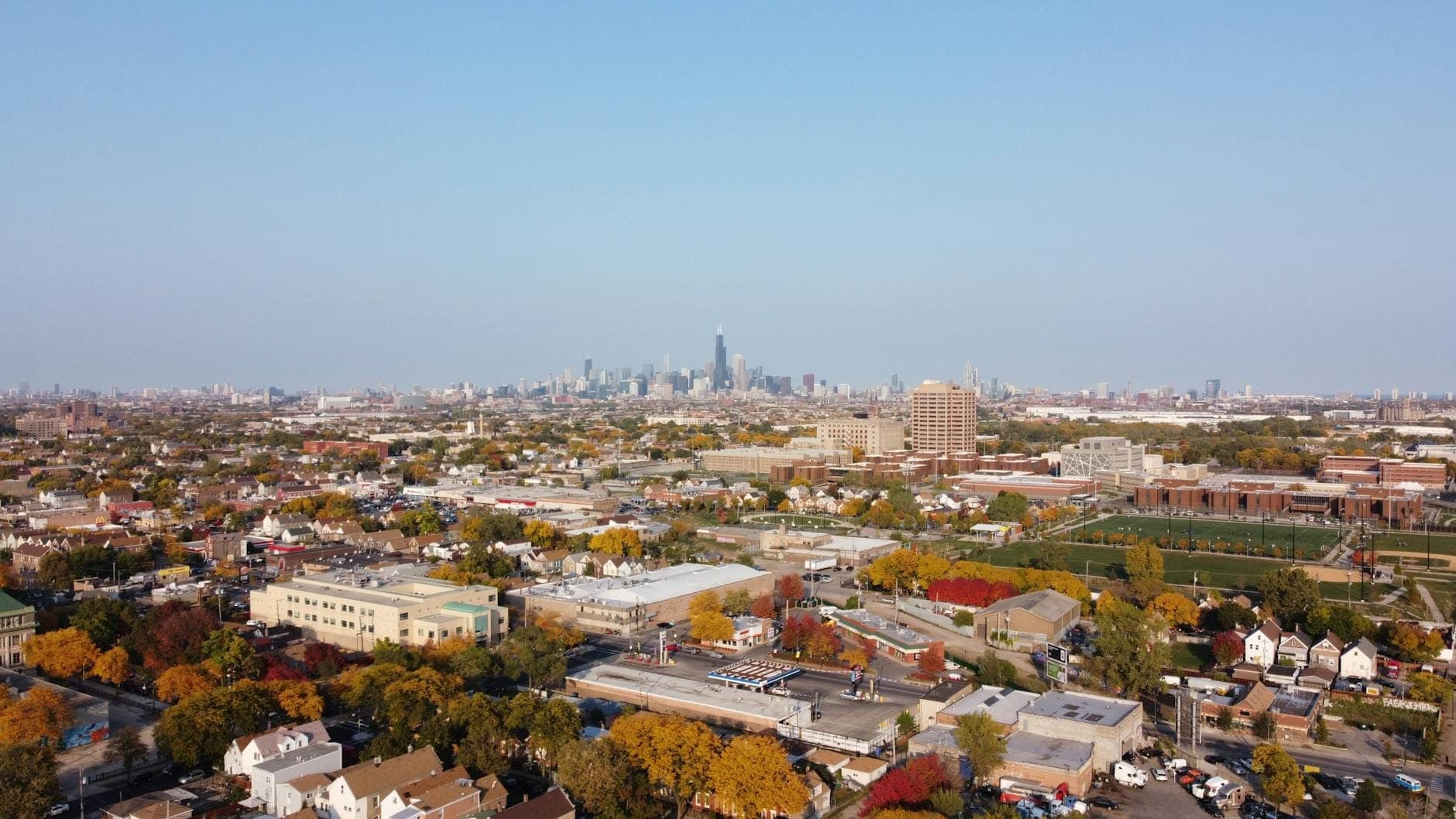 A wide aerial view of a Chicago neighborhood with houses and trees in the foreground, and the downtown Chicago skyline in the distance under a clear blue sky.