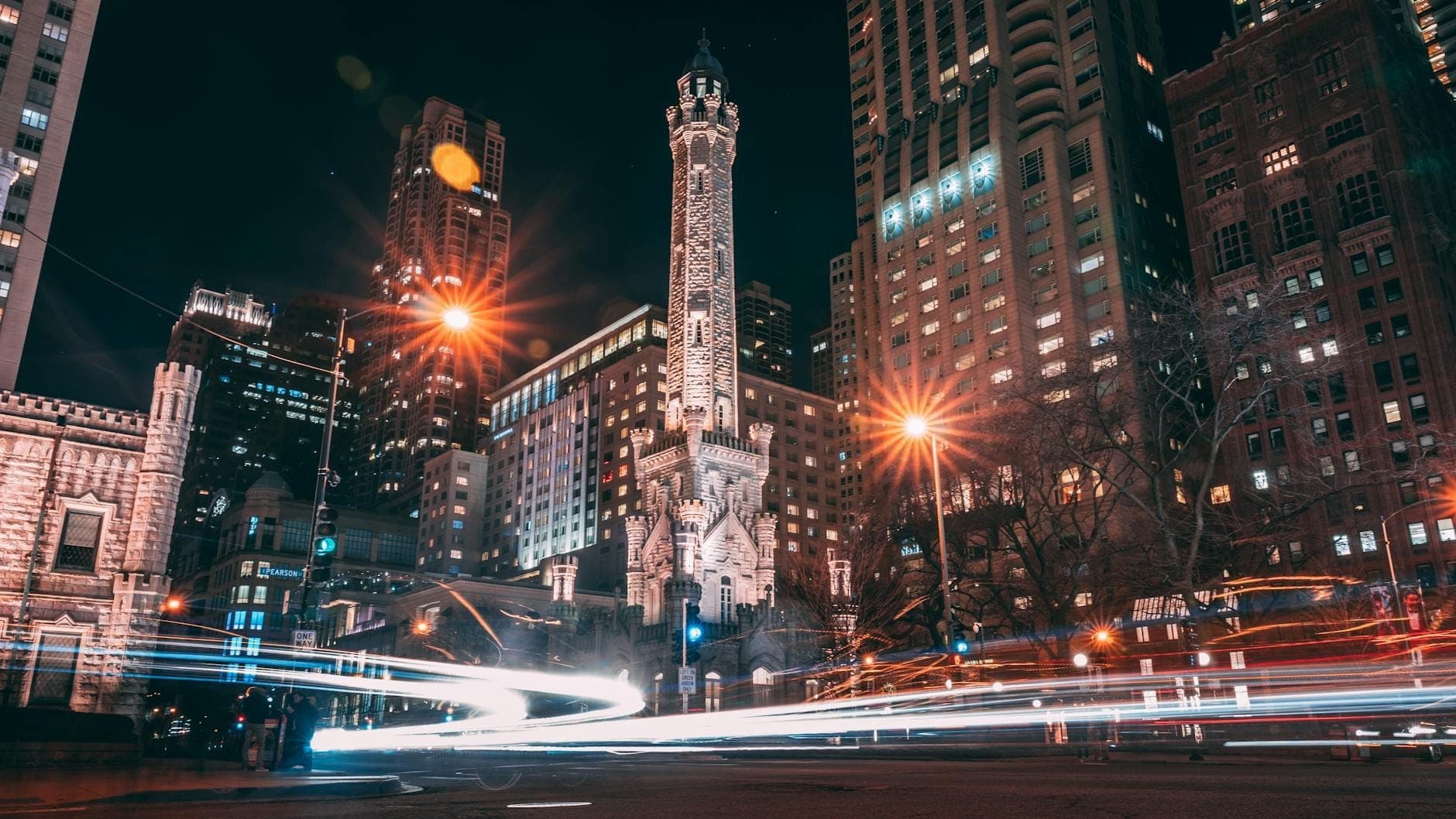 Night view of the illuminated Chicago Water Tower surrounded by modern skyscrapers on North Michigan Avenue, with light trails from cars adding dynamic energy.
