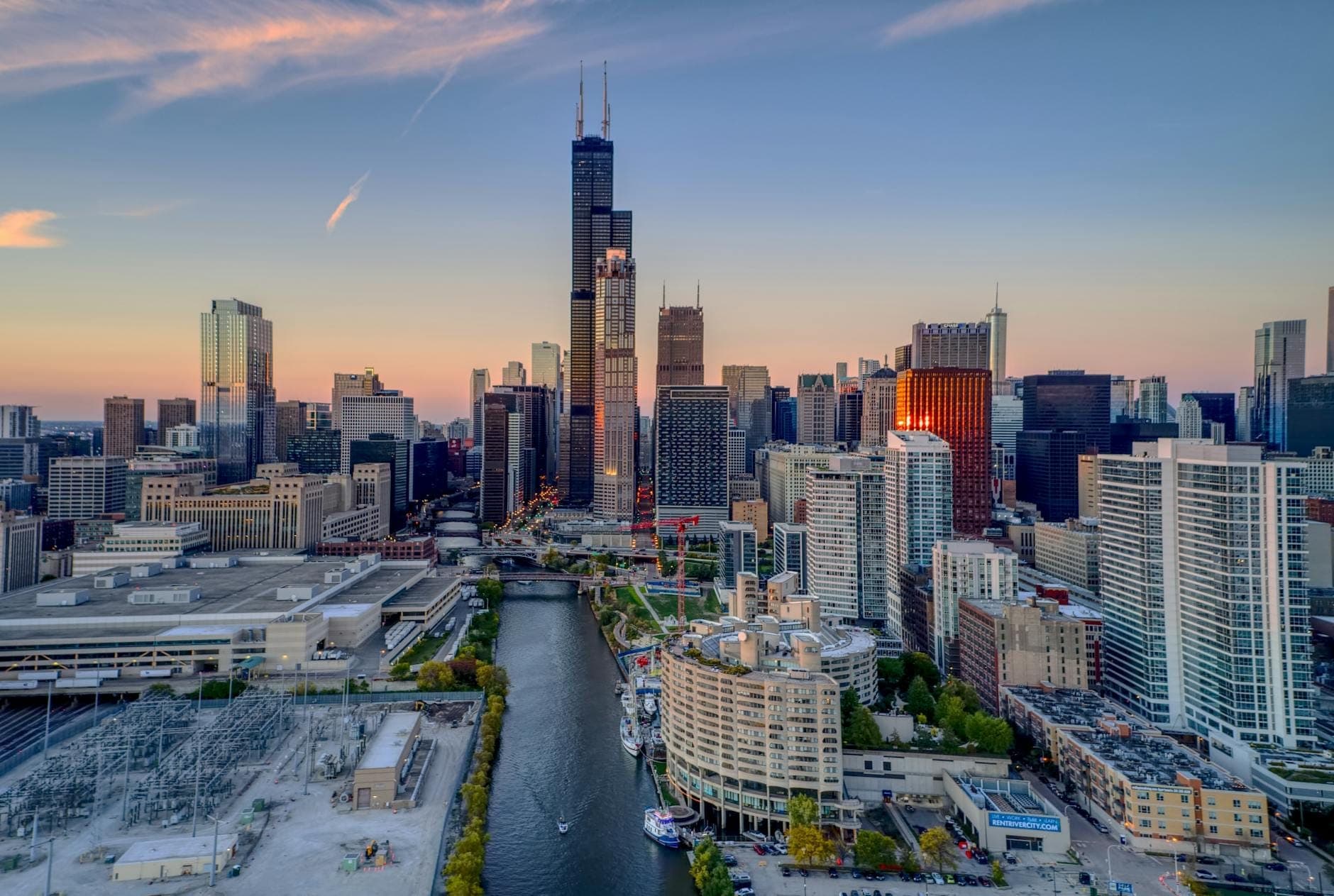 Aerial view of downtown Chicago with the river running through skyscrapers, showing distinct neighborhoods and city layout at sunset.