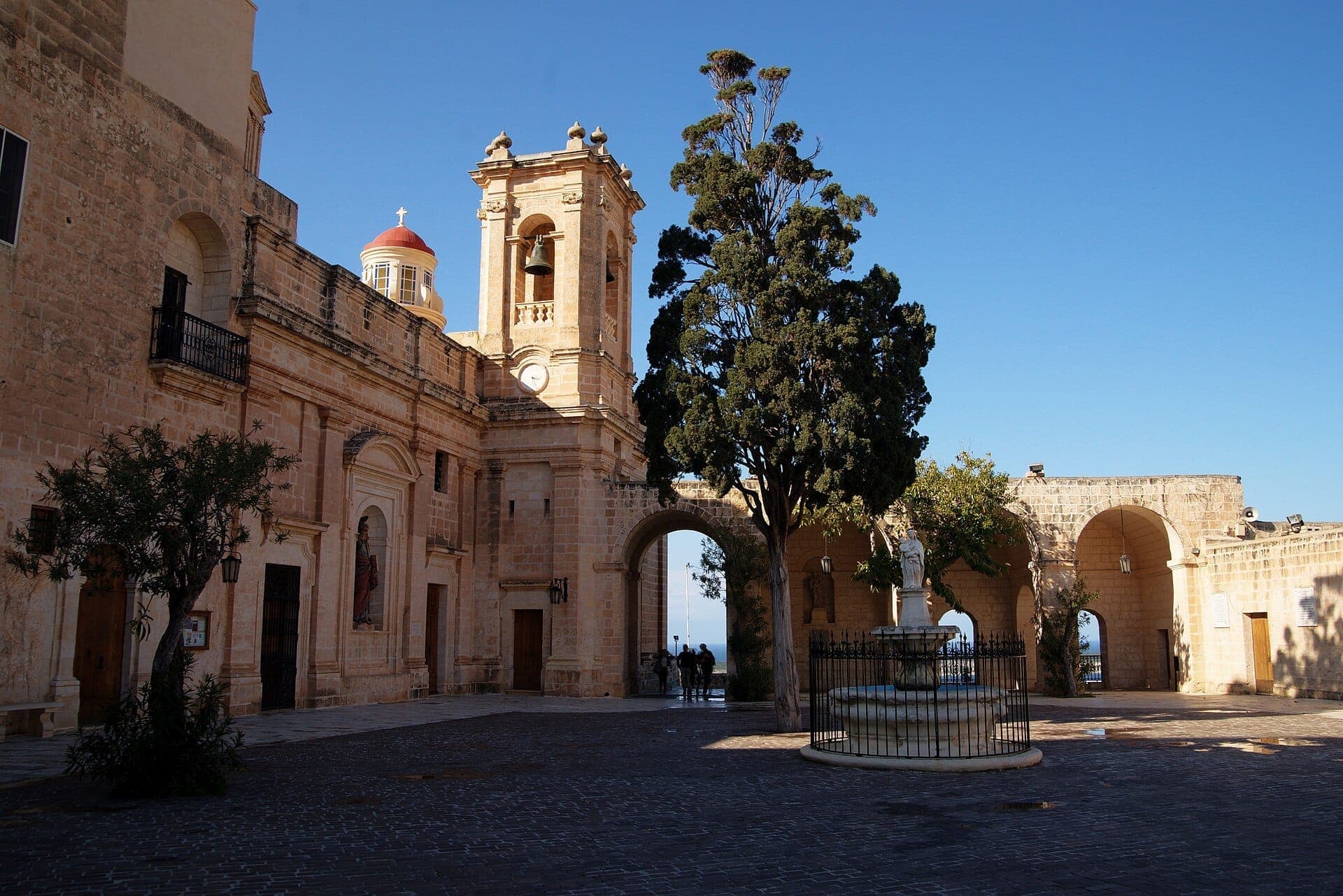 Innenhof des Heiligtums Unserer Lieben Frau von Mellieħa mit Kalksteinmauern, Glockenturm, Bogendurchgang, Bäumen, einer zentralen Statue und klarem blauen Himmel.