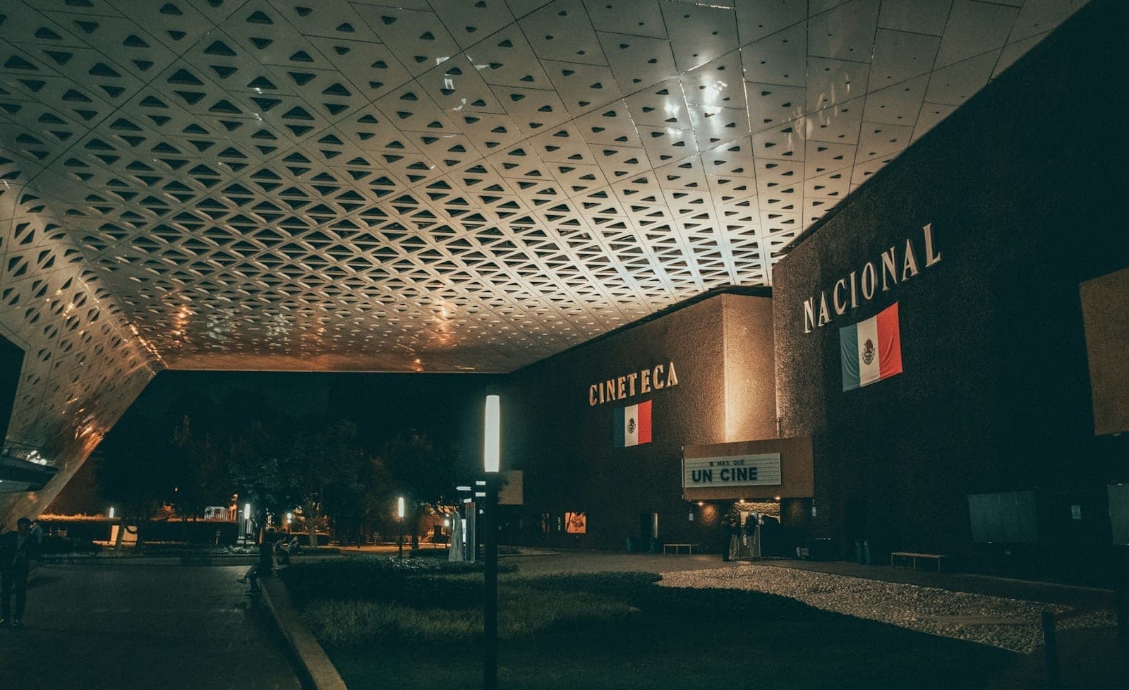 วิวยามค่ำคืนของ Cineteca Nacional ในเม็กซิโกซิตี้ โดดเด่นด้วยหลังคาทรงเรขาคณิตสมัยใหม่ ทางเข้าที่สว่างไสว และธงชาติเม็กซิโกเหนืออาคารหลัก