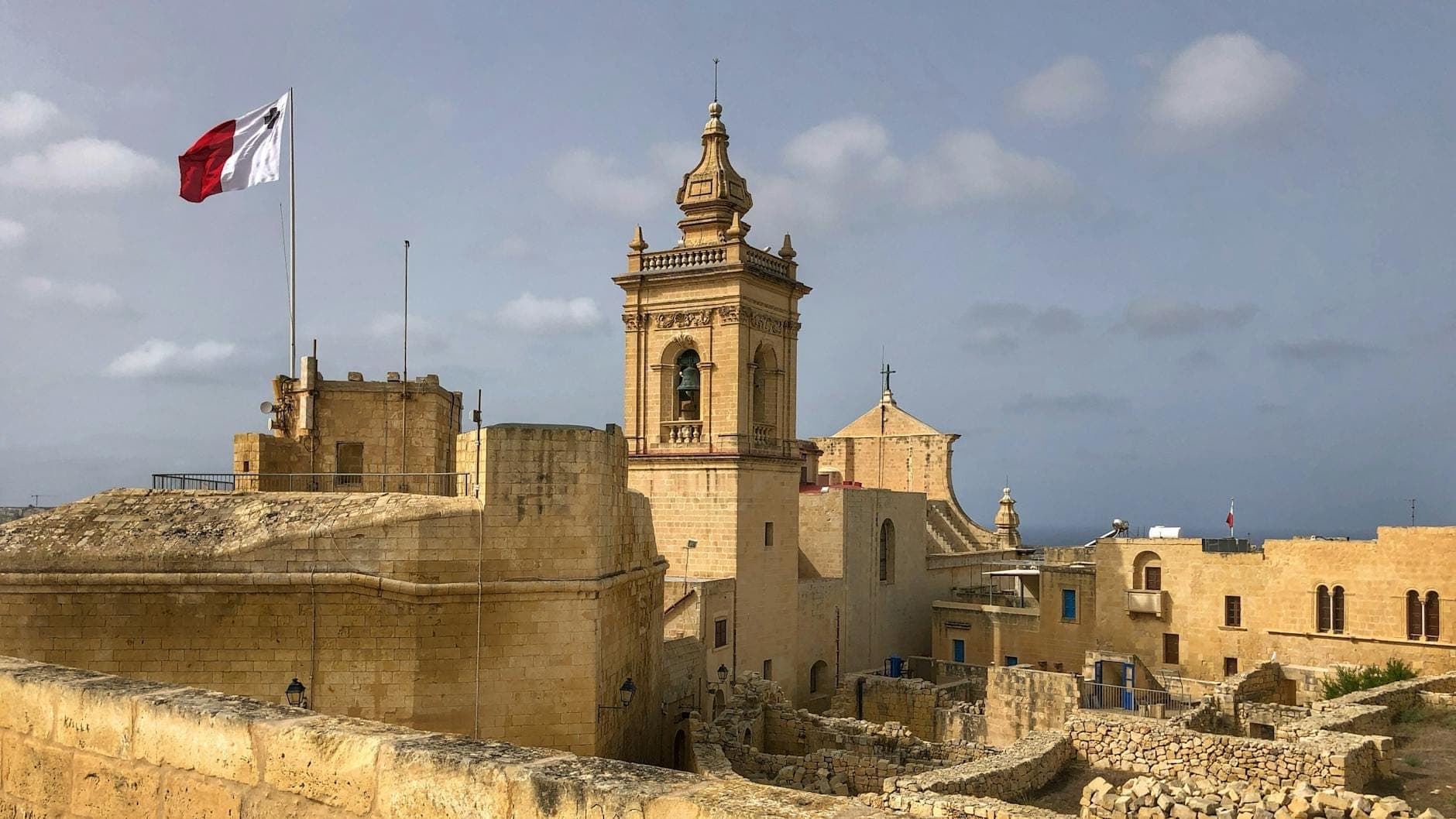 Panoramablick auf die Citadella-Festung in Victoria, Gozo, mit historischen Steinmauern, Glockenturm, Kathedrale und maltesischer Flagge unter einem teils bewölkten Himmel.