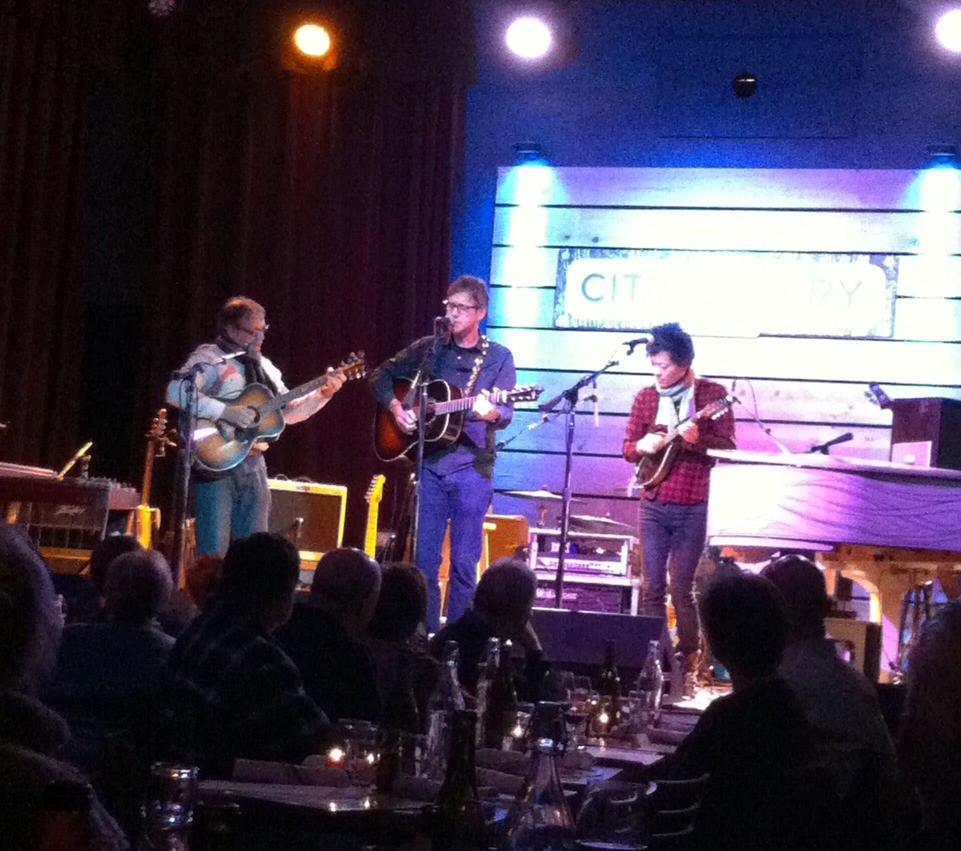 Three musicians perform with guitars and mandolin on the warmly lit stage at City Winery Nashville, while an audience sits at candlelit tables.