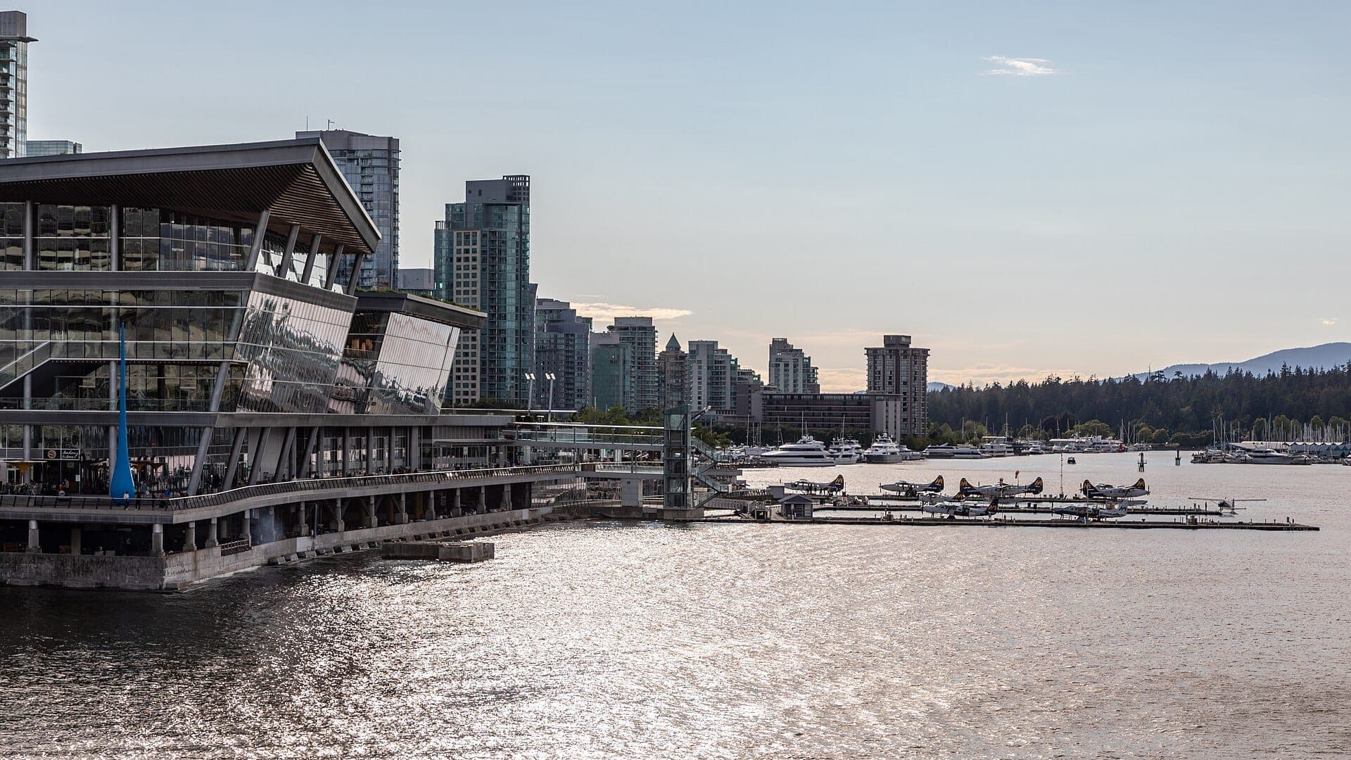 อาคารกระจกสมัยใหม่มองเห็นมารีนาและเครื่องบินทะเลที่ Coal Harbour พร้อมเส้นขอบฟ้าของ Vancouver และน้ำที่สงบนิ่งใต้แสงยามเย็นอันอ่อนโยน