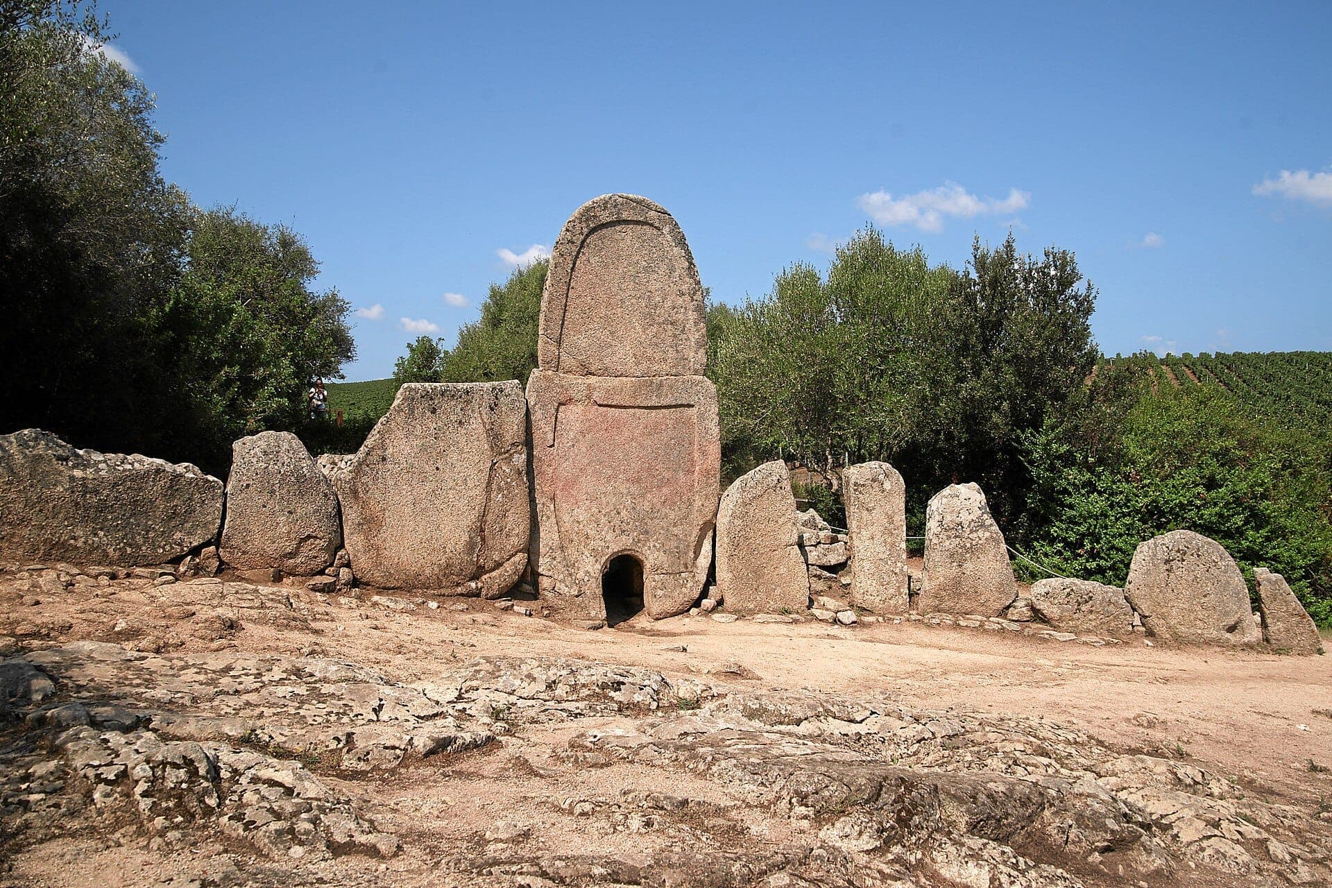 Vista frontal de la Tumba de los Gigantes de Coddu Vecchiu, con grandes piedras de granito dispuestas en semicírculo entre árboles verdes y cielo azul.