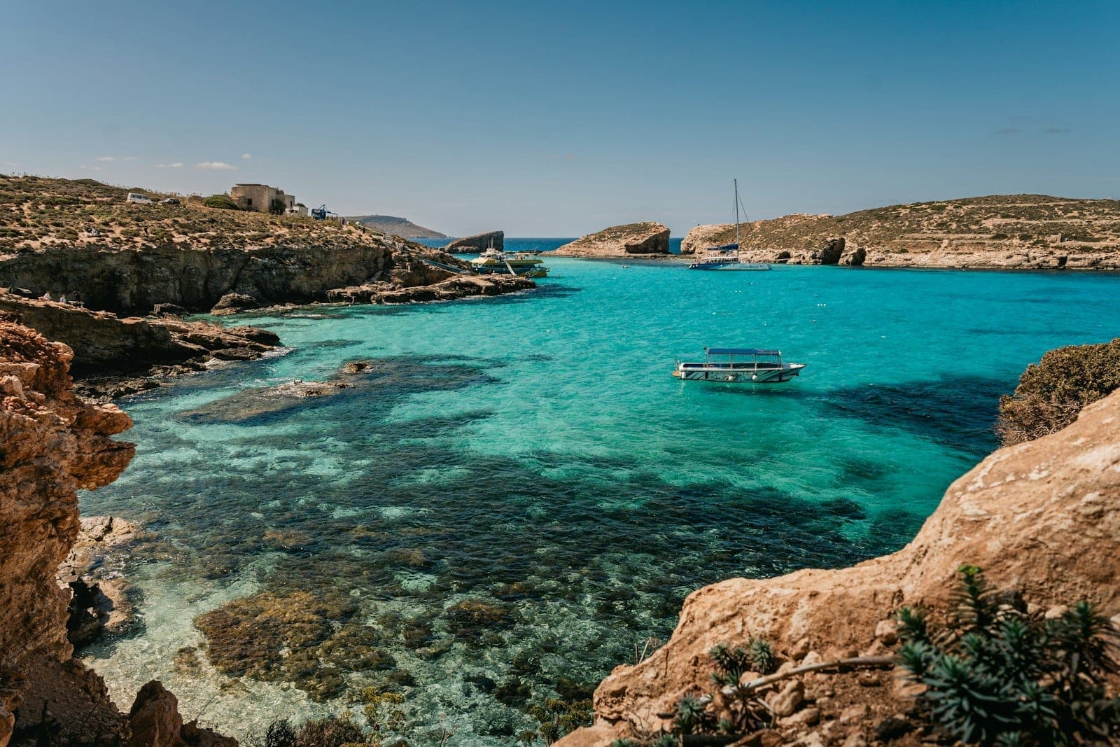 Das türkisfarbene Wasser der Blauen Lagune von Comino, umgeben von felsigen Küsten unter strahlend blauem Himmel, mit einer schwimmenden Plattform in der Lagune.