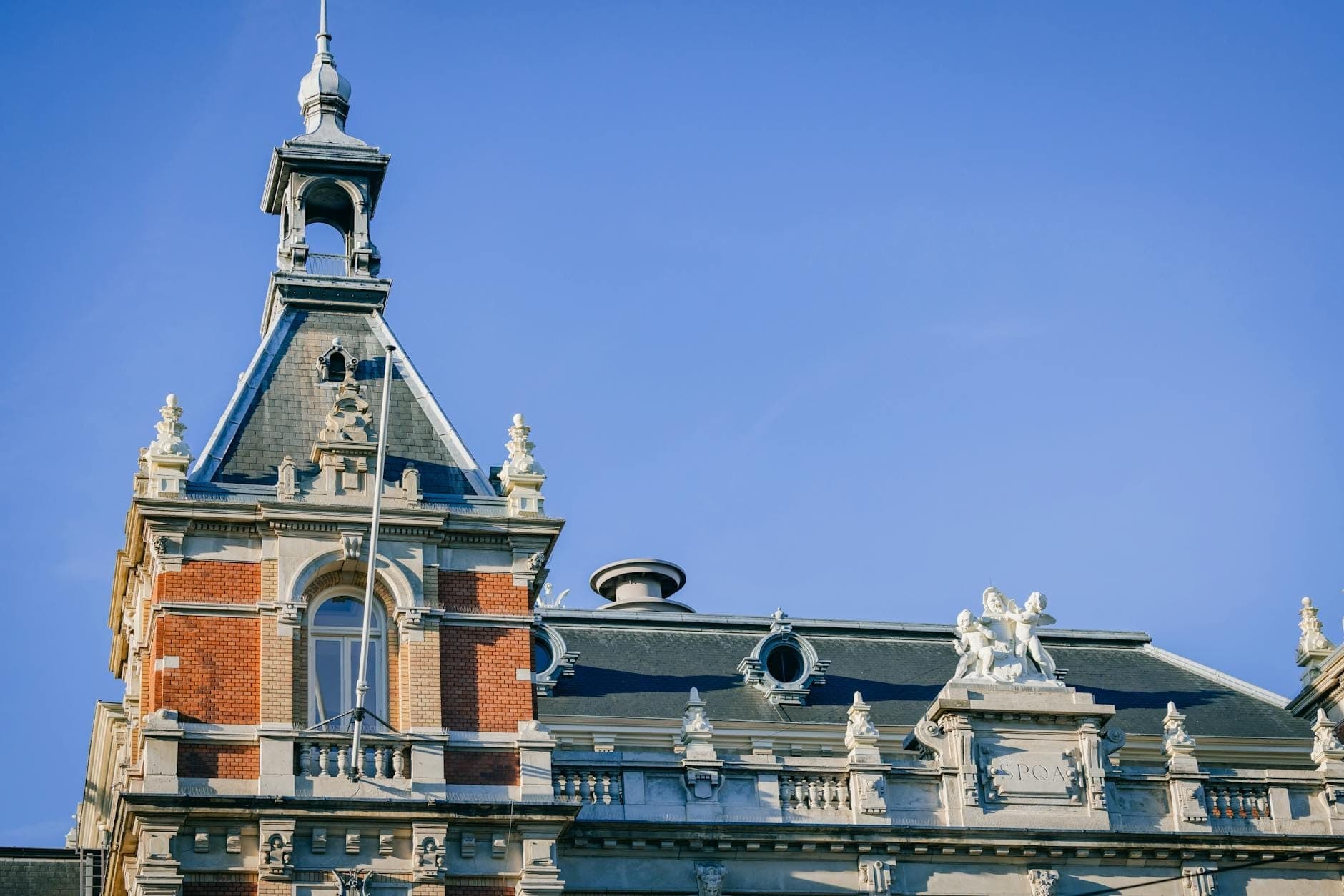 Prachtvoller oberer Abschluss eines eleganten Konzertsaalgebäudes in Amsterdam mit dekorativen Statuen, aufwendiger Architektur und klarem blauem Himmel im Hintergrund.