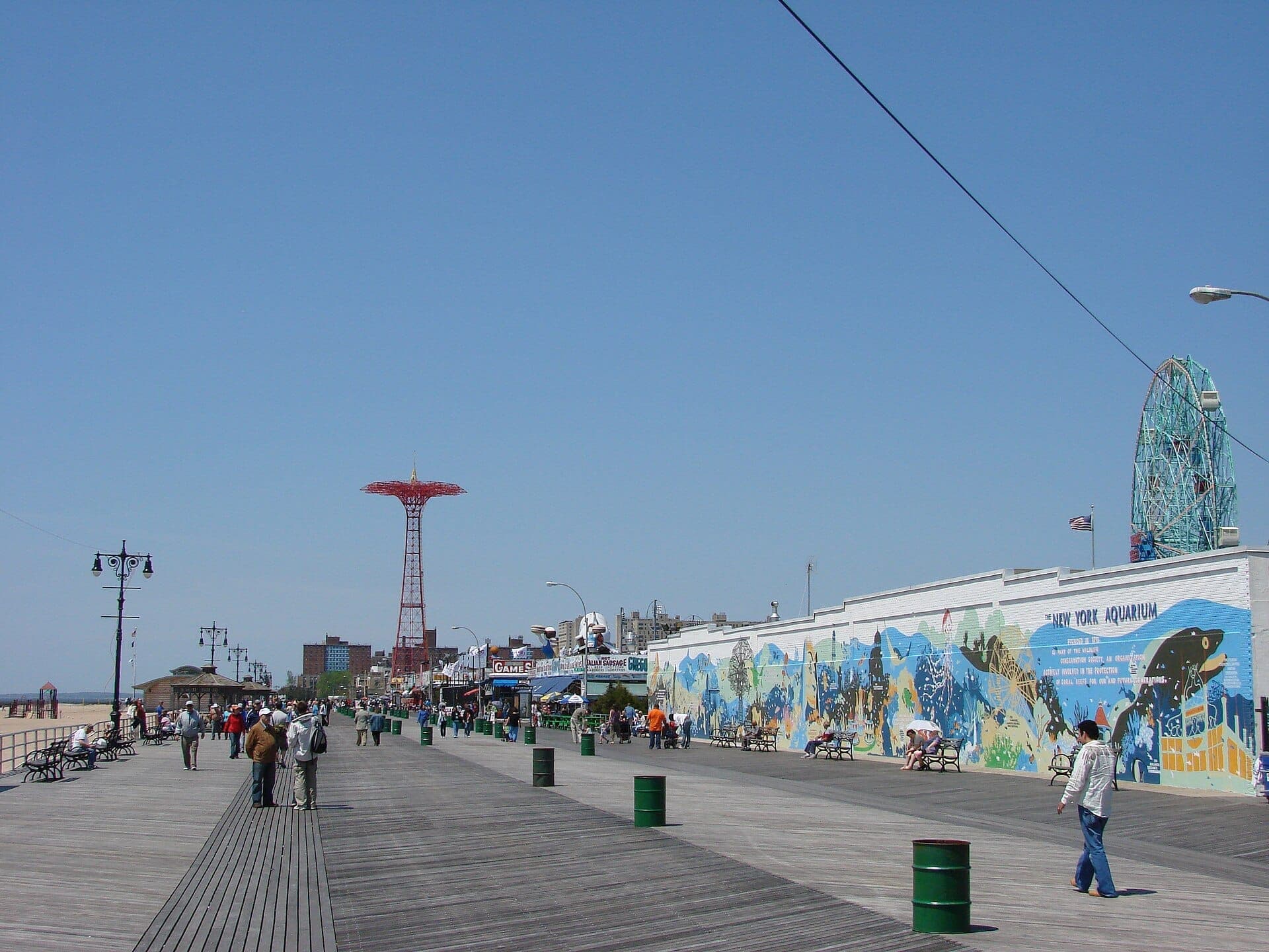 นักท่องเที่ยวเดินเล่นตามแนว Coney Island Boardwalk ท่ามกลางท้องฟ้าสีฟ้า มีภาพจิตรกรรมฝาผนังบนกำแพงพิพิธภัณฑ์สัตว์น้ำ หอคอย Parachute Jump และ Wonder Wheel อยู่เบื้องหลัง