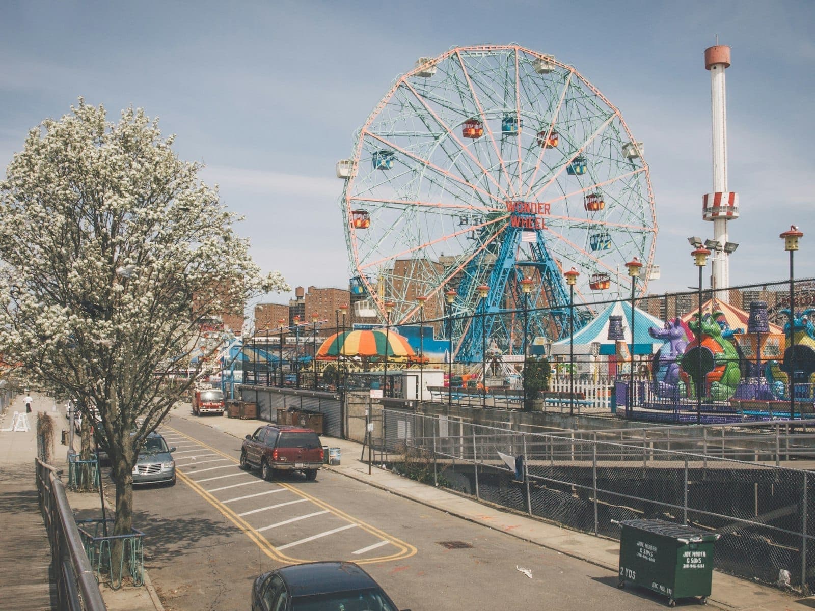 ภาพมุมกว้างสวนสนุกโคนีย์ไอแลนด์ เห็นชิงช้าสวรรค์ Wonder Wheel ต้นไม้ผลิบาน และเครื่องเล่นสีสันสดใสในวันที่แดดจ้า