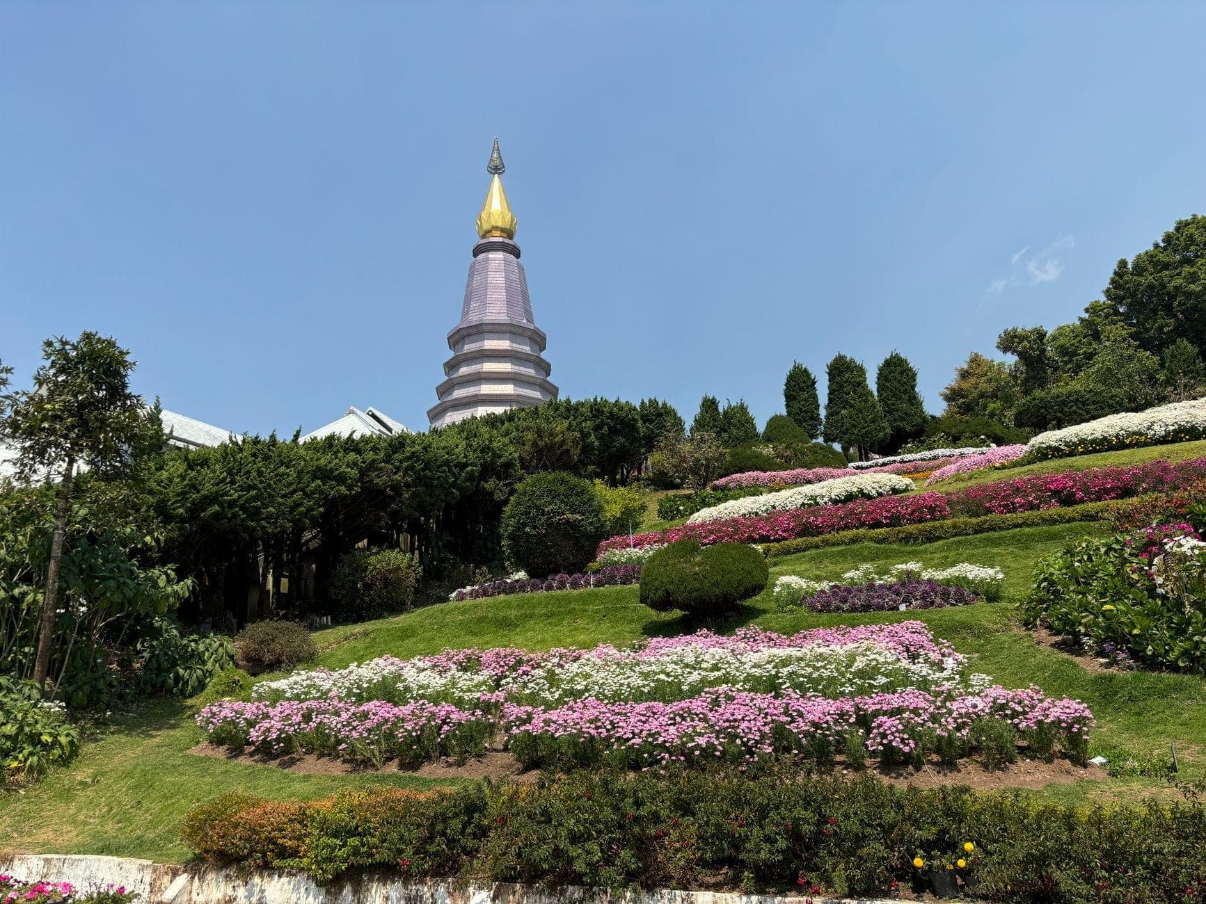 Un rigoglioso giardino collinare con aiuole di fiori colorati e il caratteristico Chedi dalla sommità viola del tempio Doi Inthanon che si eleva tra il verde a Chiang Mai.