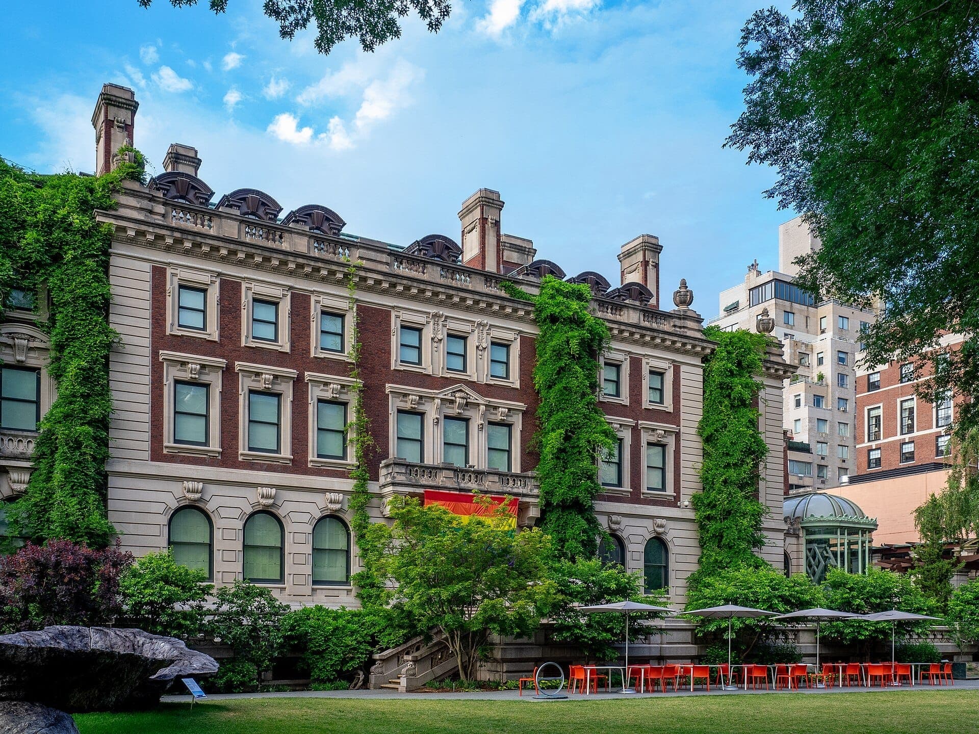 ภายนอกของ Cooper Hewitt, Smithsonian Design Museum มีคฤหาสน์อิฐประวัติศาสตร์ที่ปกคลุมด้วยไม้เลื้อย พร้อมสนามหญ้า ต้นไม้ และพื้นที่นั่งเล่นกลางแจ้ง