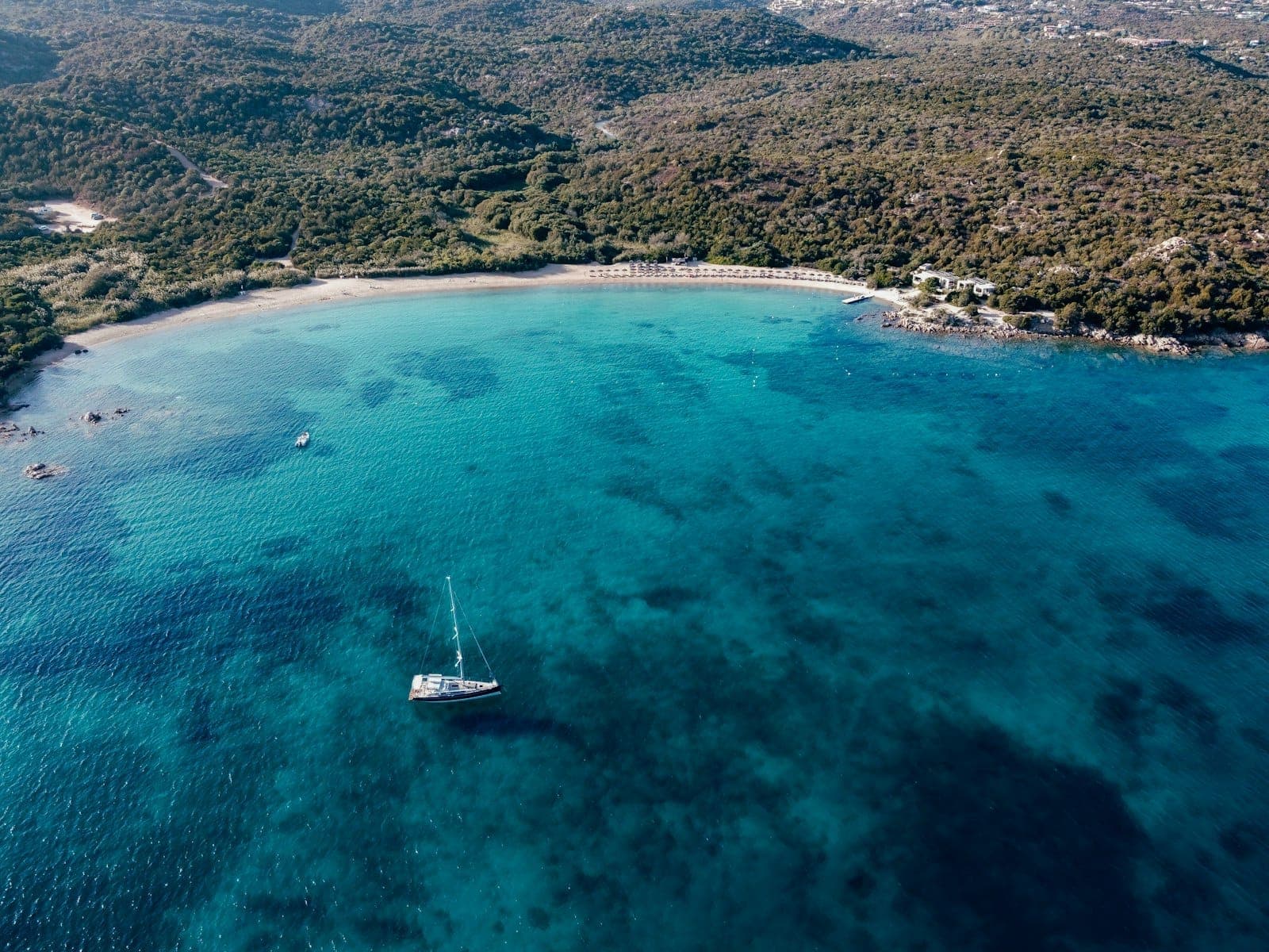 Vista aérea de una bahía esmeralda con playa de arena blanca, colinas verdes exuberantes y un yate anclado en las cristalinas aguas turquesas de la Costa Esmeralda.