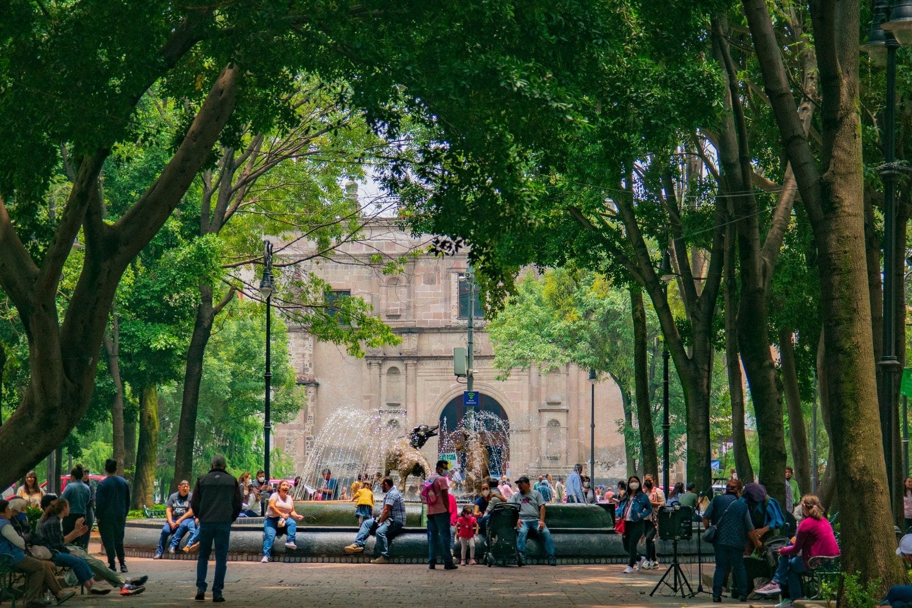 นักท่องเที่ยวรวมตัวกันรอบน้ำพุกลางจัตุรัสหลักของ Coyoacán ล้อมรอบด้วยต้นไม้สีเขียวสูงใหญ่และด้านหน้าโบสถ์หินที่มีประวัติศาสตร์