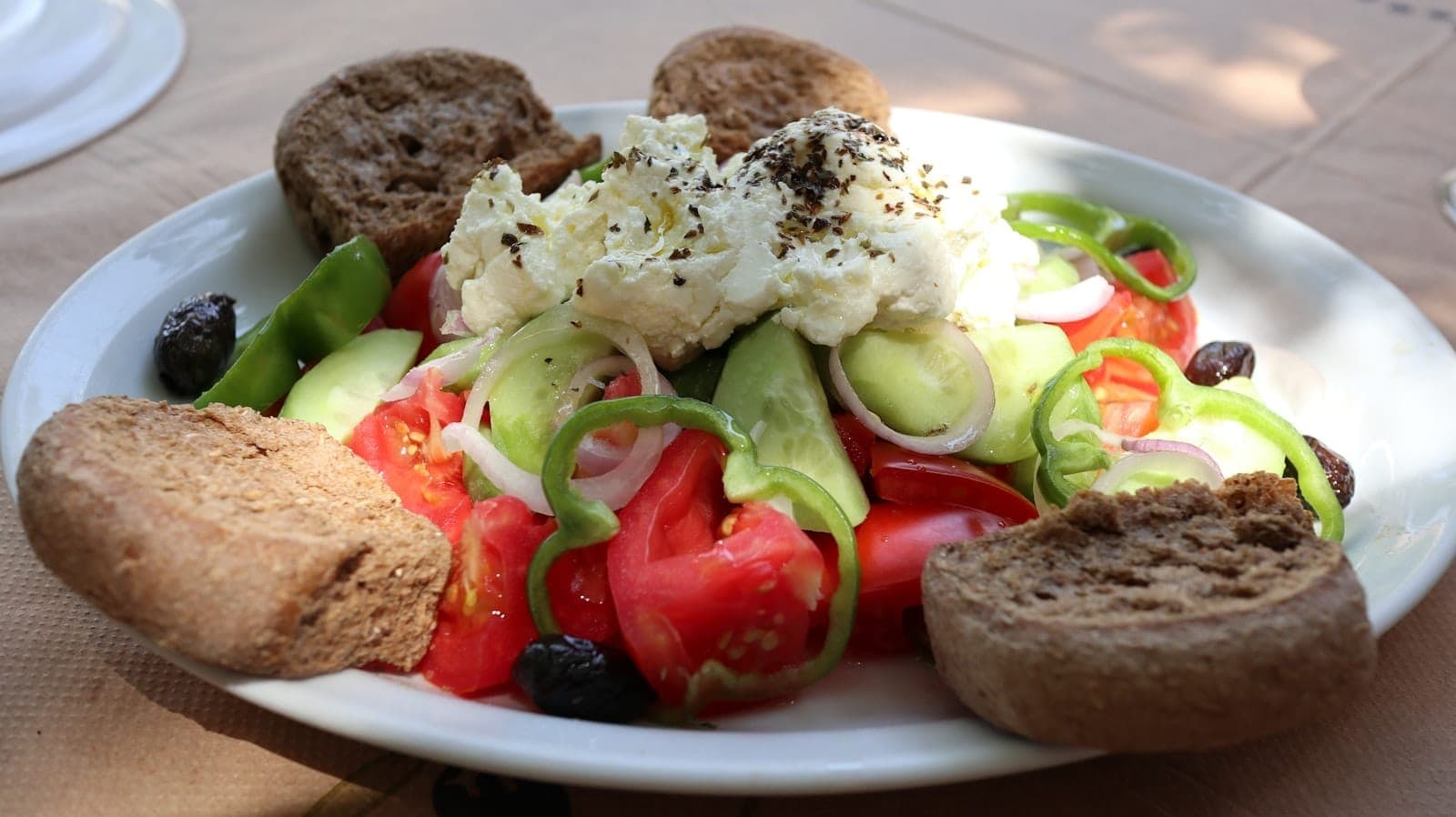 Ein Teller traditioneller kretischer Dakos-Salat mit Tomaten, Gurken, grünen Paprika, Oliven und cremigem Käse, umgeben von Gerstenbroten, auf einem sonnenbeschienenen Tisch im Freien.