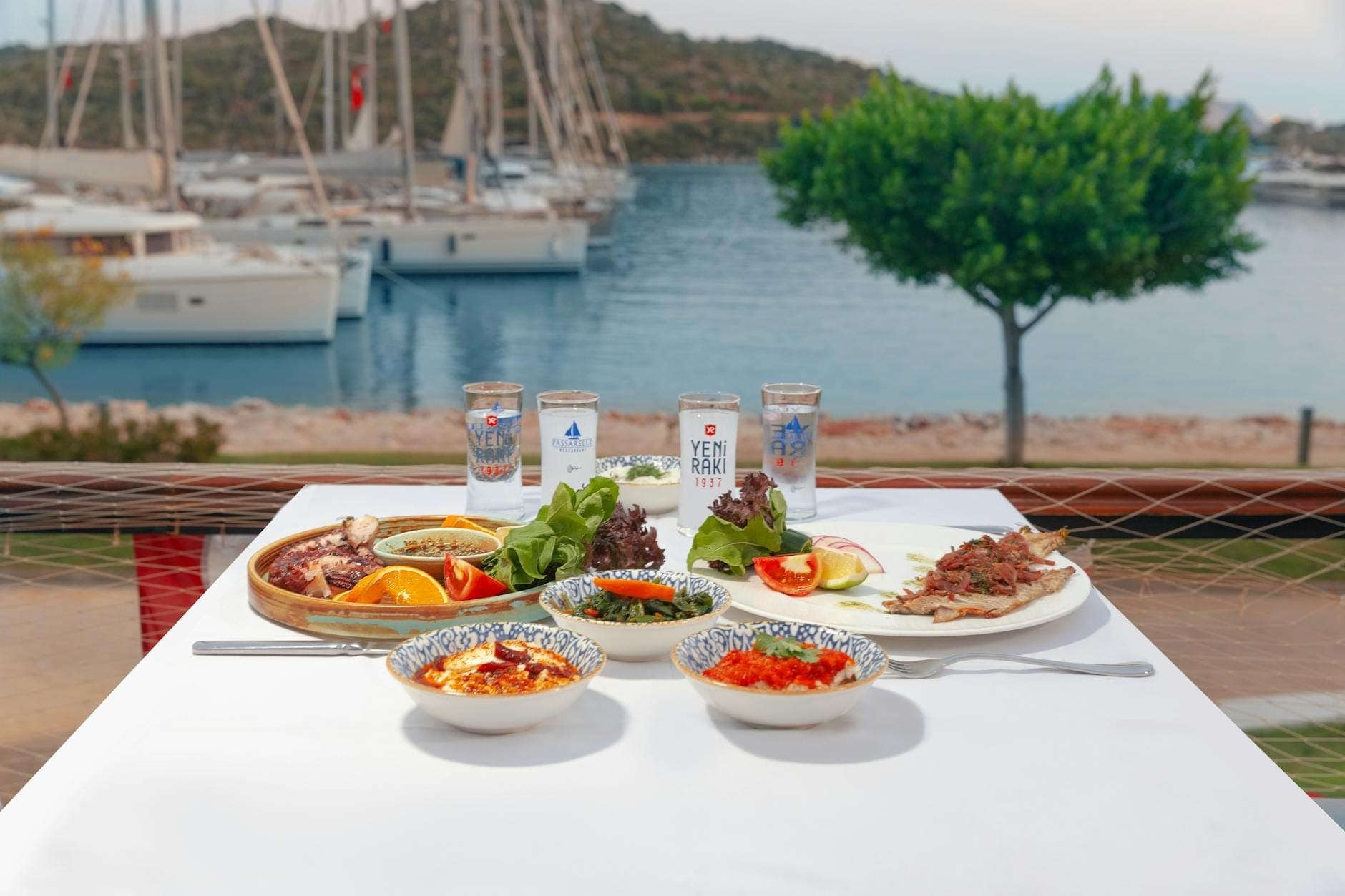 Gedeckter Tisch mit kretischen Gerichten, Raki-Gläsern und Blick auf einen Yachthafen mit Booten und Hügeln im Hintergrund.