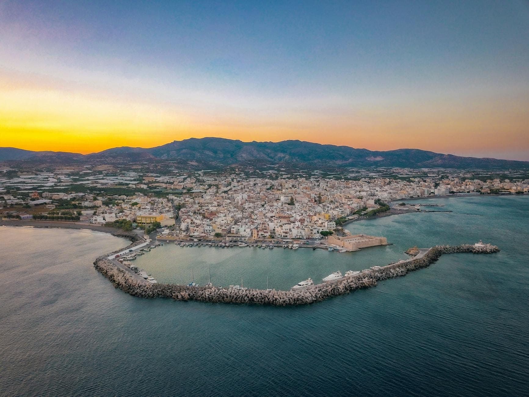 Aerial view of the coastal city of Rethymno in Crete at sunset, showing the picturesque harbor, mountains, and waterfront buildings.