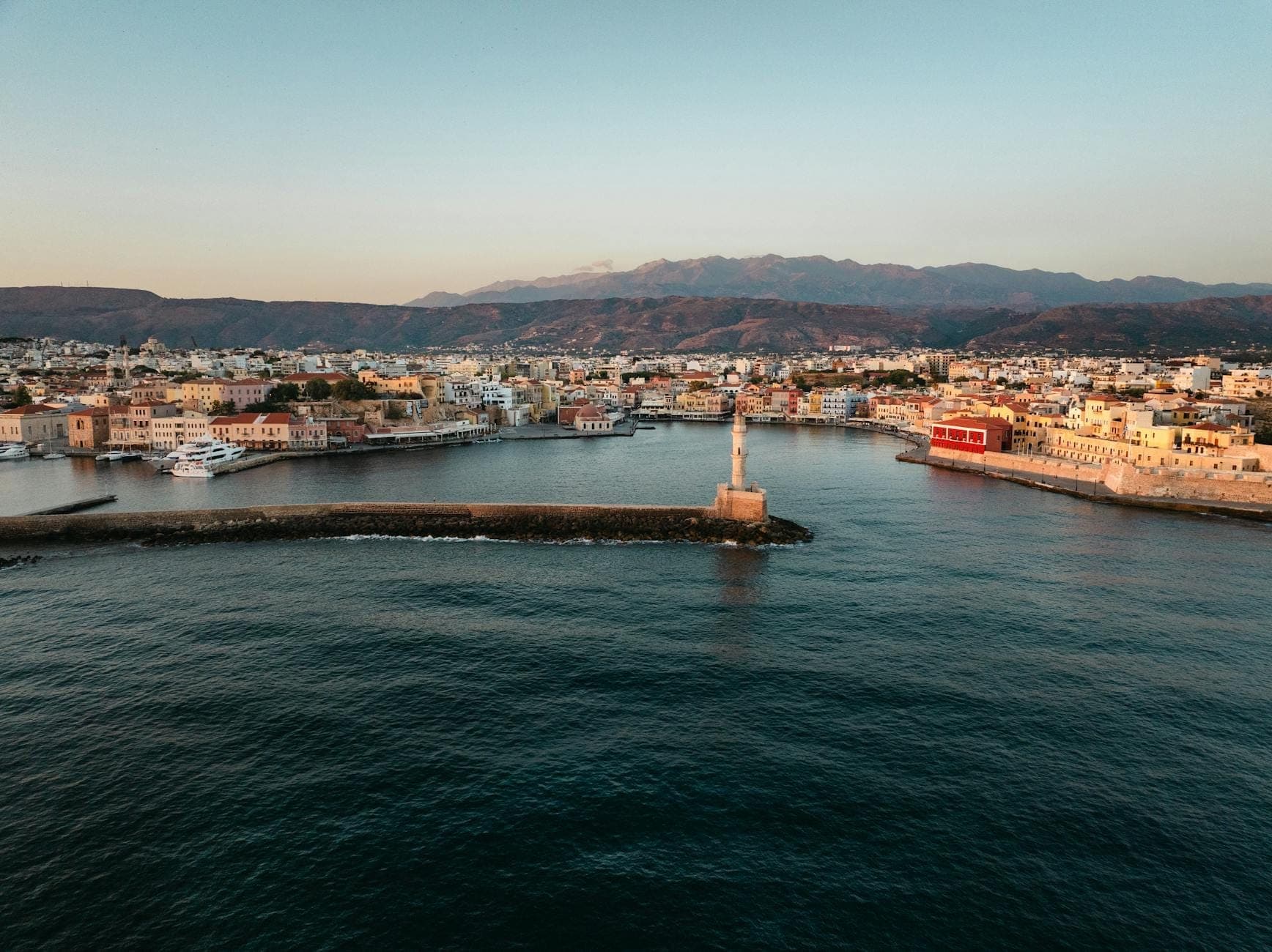 Vista aerea del porto veneziano di Chania, Creta, con un faro, edifici sul lungomare e montagne sullo sfondo.