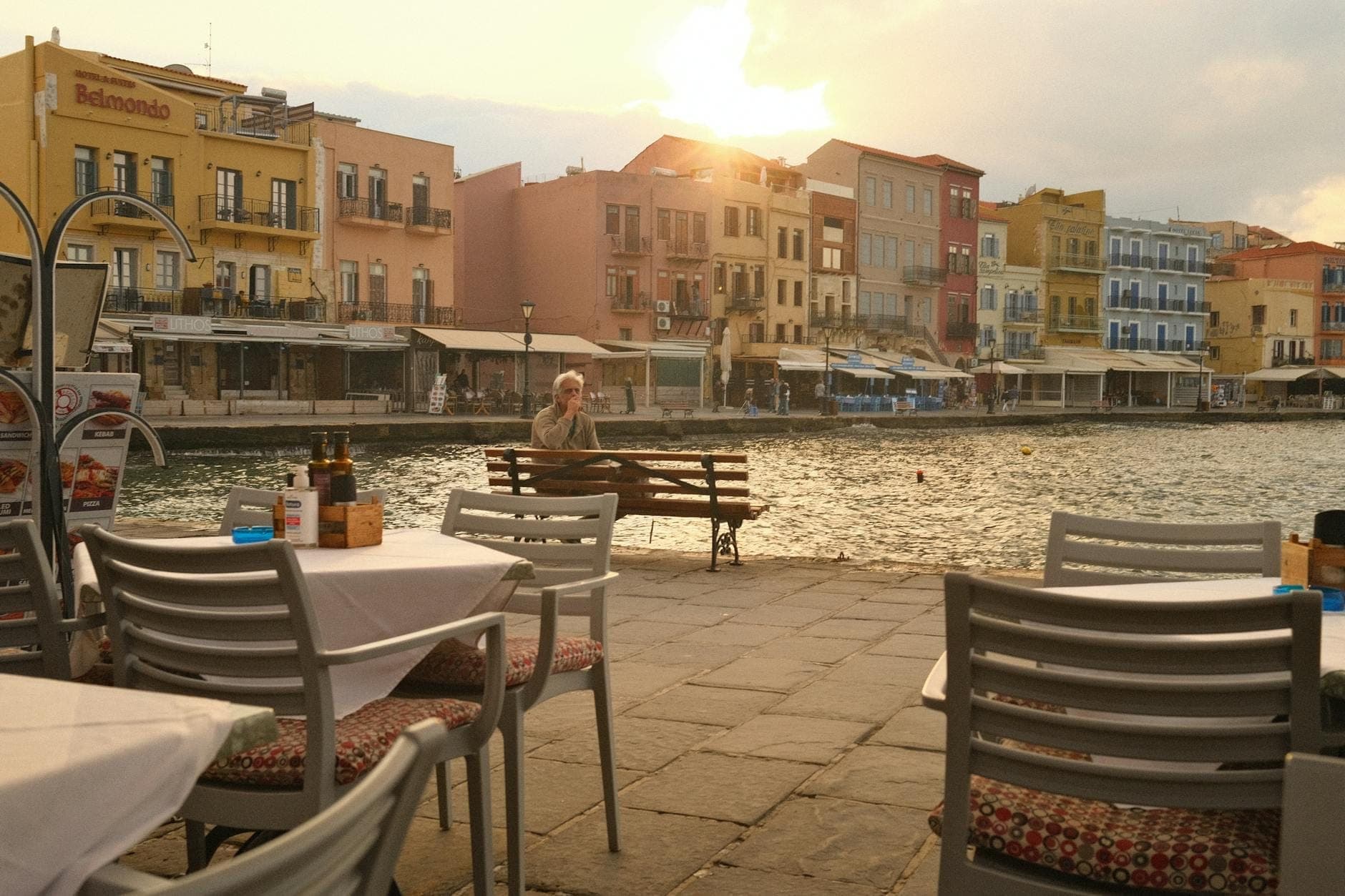 Tavoli vuoti di ristorante all'aperto al tramonto nel Porto Veneziano di Creta, con edifici colorati e acqua sullo sfondo.