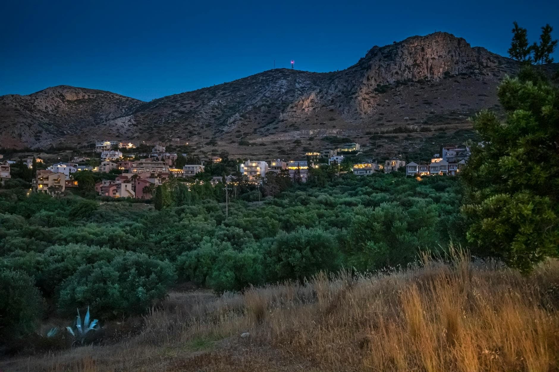 Bergdorf auf Kreta in der Abenddämmerung, mit Häusern zwischen Olivenhainen und schroffen Hügeln unter einem tiefdunklen Zwielicht.