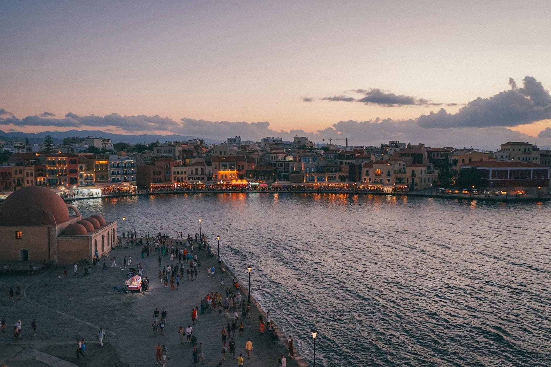 Abendlicher Blick auf den venezianischen Hafen von Chania auf Kreta, mit Altstadtgebäuden, Uferpromenade und Menschenmassen, die bei Sonnenuntergang die lebhafte Atmosphäre genießen.