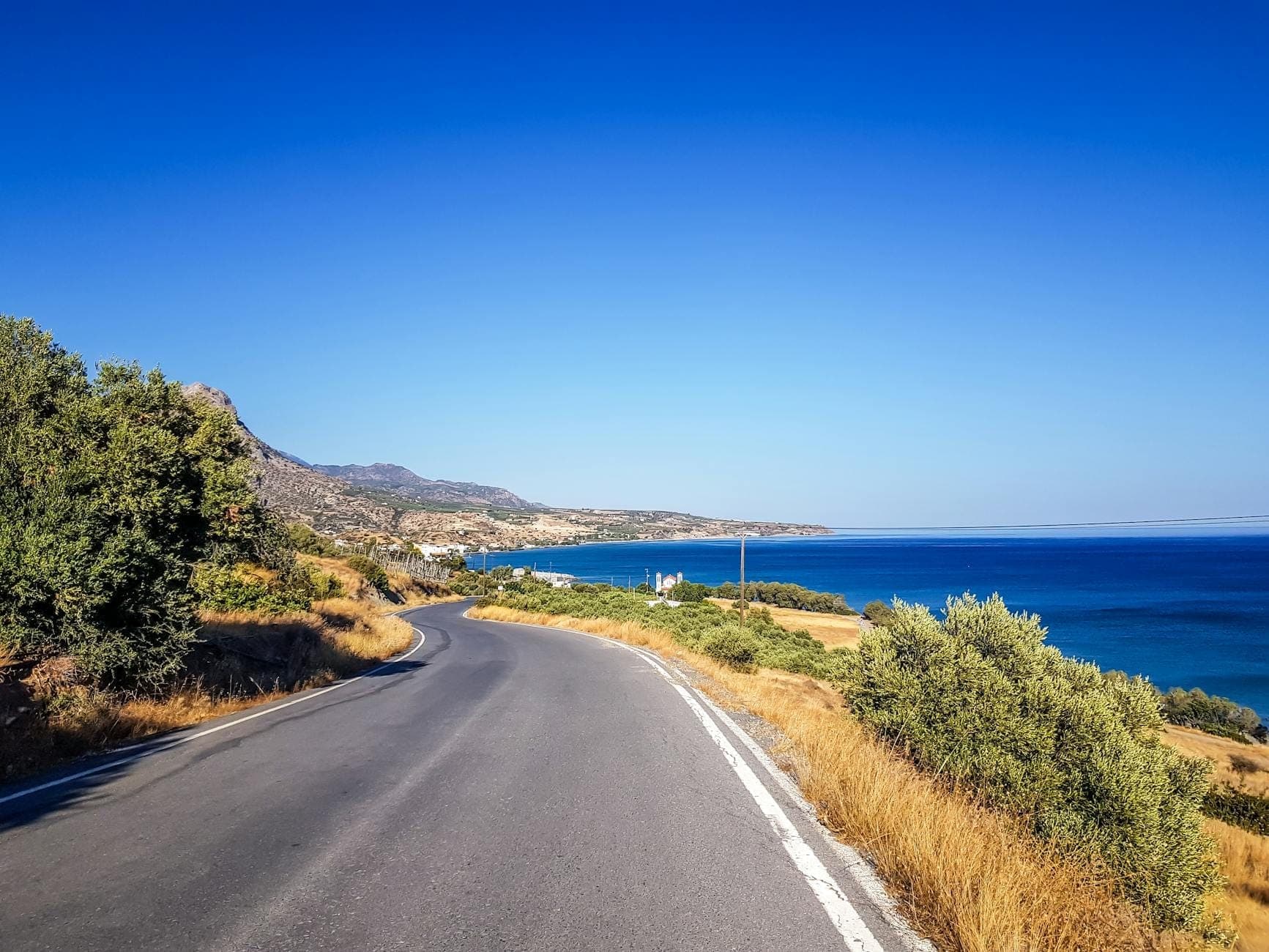 Kurvenreiche Küstenstraße auf Kreta mit blauem Meer auf der rechten Seite, goldenem Gras und Hügeln unter einem klaren blauen Himmel.
