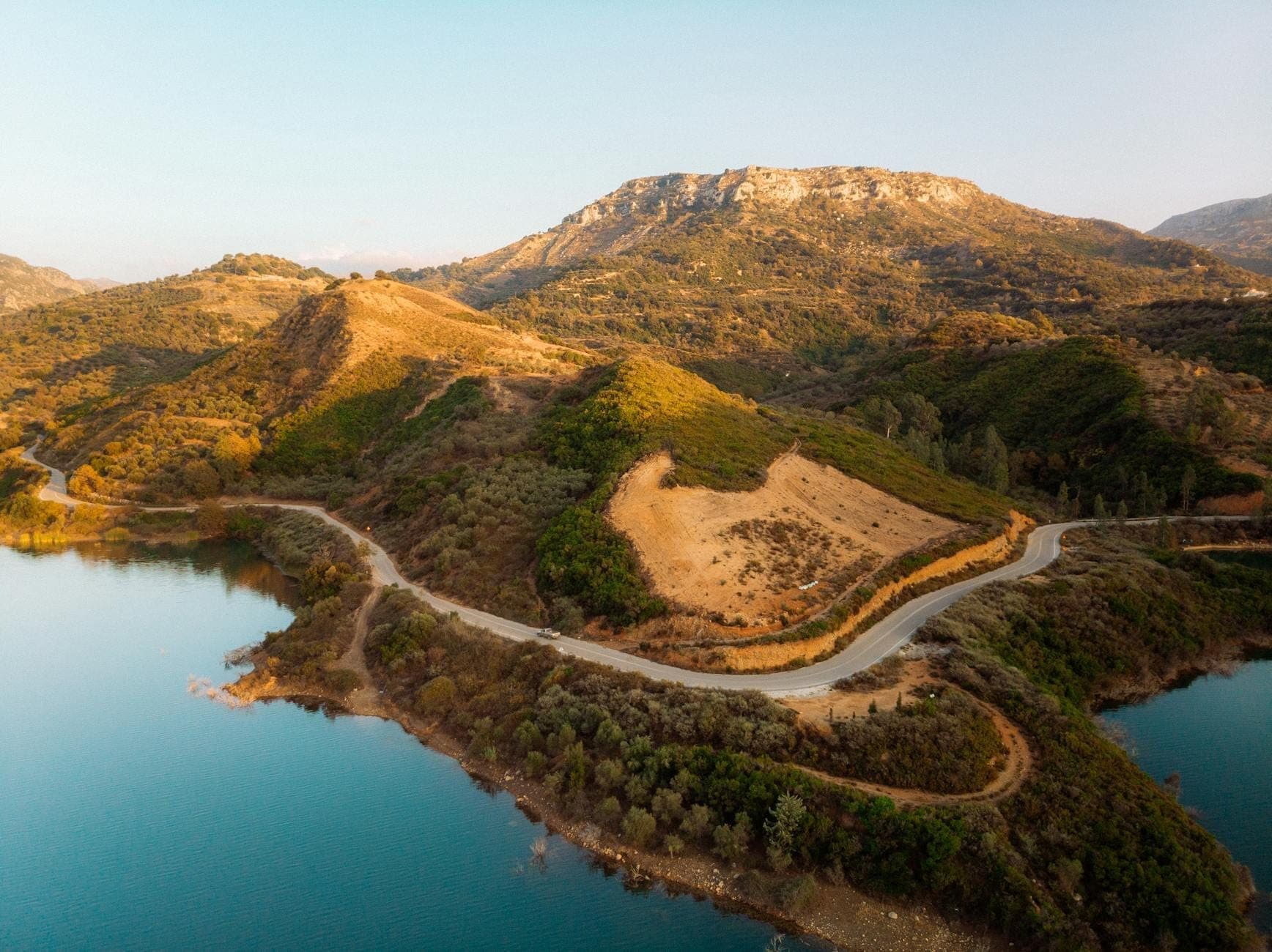 Kurvenreiche Küstenstraße auf Kreta mit türkisfarbenem Wasser, üppigen Hügeln und einer bergigen Kulisse unter klarem Himmel.